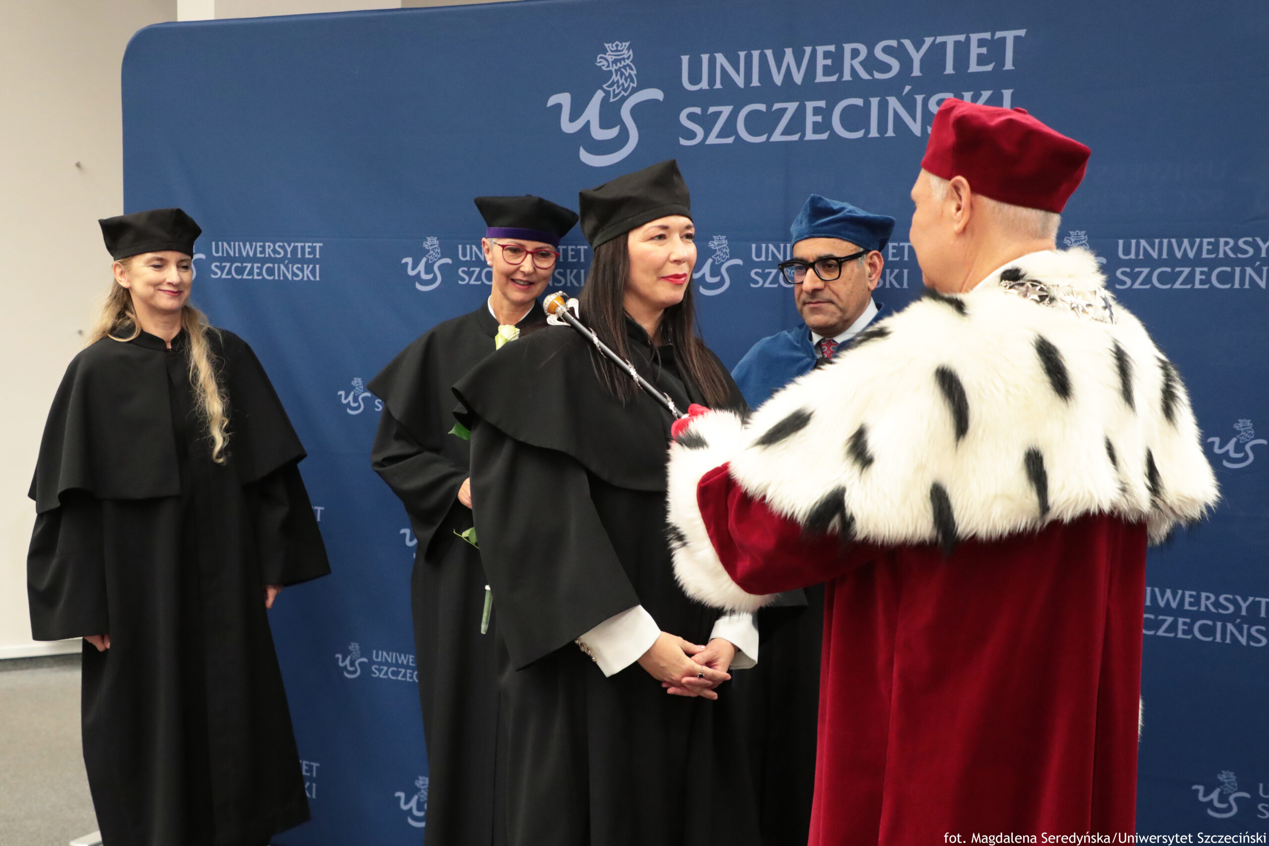 Ceremonia akademicka na tle niebieskiej ścianki z napisem „UNIWERSYTET SZCZECIŃSKI” i logo „US”. Na pierwszym planie stoi kobieta w czarnej todze i birecie, z długimi ciemnymi włosami, jasną skórą i czerwonymi ustami; trzyma ręce złożone z przodu. Z prawej strony, tyłem do aparatu, stoi mężczyzna w czerwonej, uroczystej szacie z białym futrzanym kołnierzem w czarne plamy oraz w czerwonym nakryciu głowy; trzyma w ręku berło lub laskę ceremonialną, skierowaną w stronę kobiety. W tle stoją jeszcze trzy osoby w togach: kobieta po lewej w czarnej todze i birecie, z długimi jasnymi włosami i jasną skórą; kobieta pośrodku z okularami, w czarnej todze i birecie; oraz mężczyzna po prawej w niebieskiej todze i birecie, w okularach, z jasną skórą. W prawym dolnym rogu widoczny jest drobny podpis: „fot. Magdalena Seredyńska/Uniwersytet Szczeciński”.