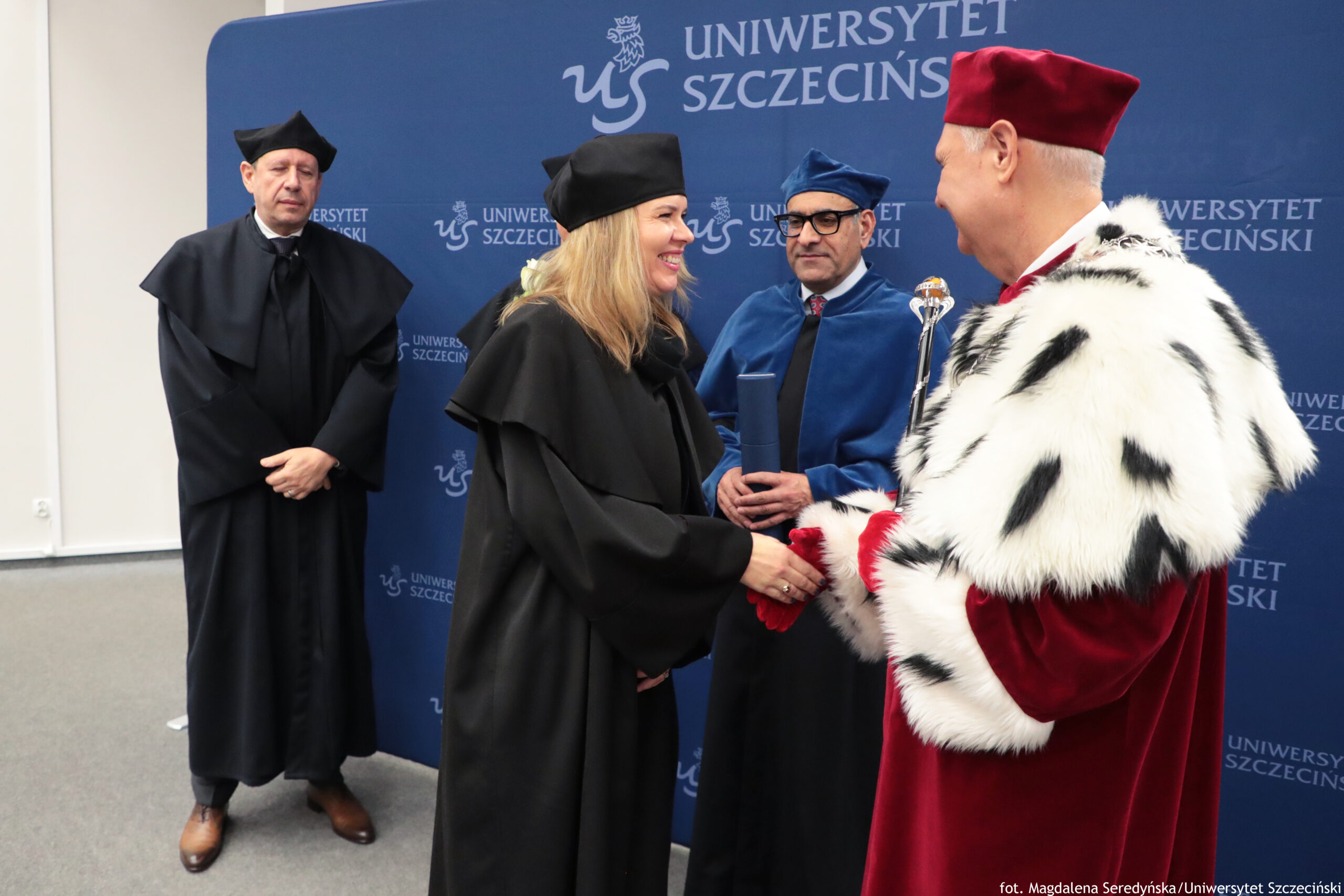 Ceremonia akademicka z czterema osobami w togach na tle ścianki z napisem „UNIWERSYTET SZCZECIŃSKI” i logo uczelni. Kobieta w czarnej todze i czarnym birecie, z blond włosami, stoi na środku i uśmiechnięta podaje dłoń mężczyźnie po prawej w czerwonym stroju ceremonialnym z białym futrzanym płaszczem w czarne plamy; mężczyzna ma czerwony biret i trzyma ozdobne berło. Za kobietą stoi mężczyzna w niebieskiej todze i niebieskim birecie, w okularach, trzyma w rękach zwinięty dokument lub dyplom. Po lewej stronie, nieco z tyłu, stoi drugi mężczyzna w czarnej todze i czarnym birecie, z rękami złożonymi z przodu. Wszyscy wyglądają na białych; scena jest wewnątrz, na szarej wykładzinie. W prawym dolnym rogu widoczny podpis fotograficzny: „fot. Magdalena Seredyńska/Uniwersytet Szczeciński”.