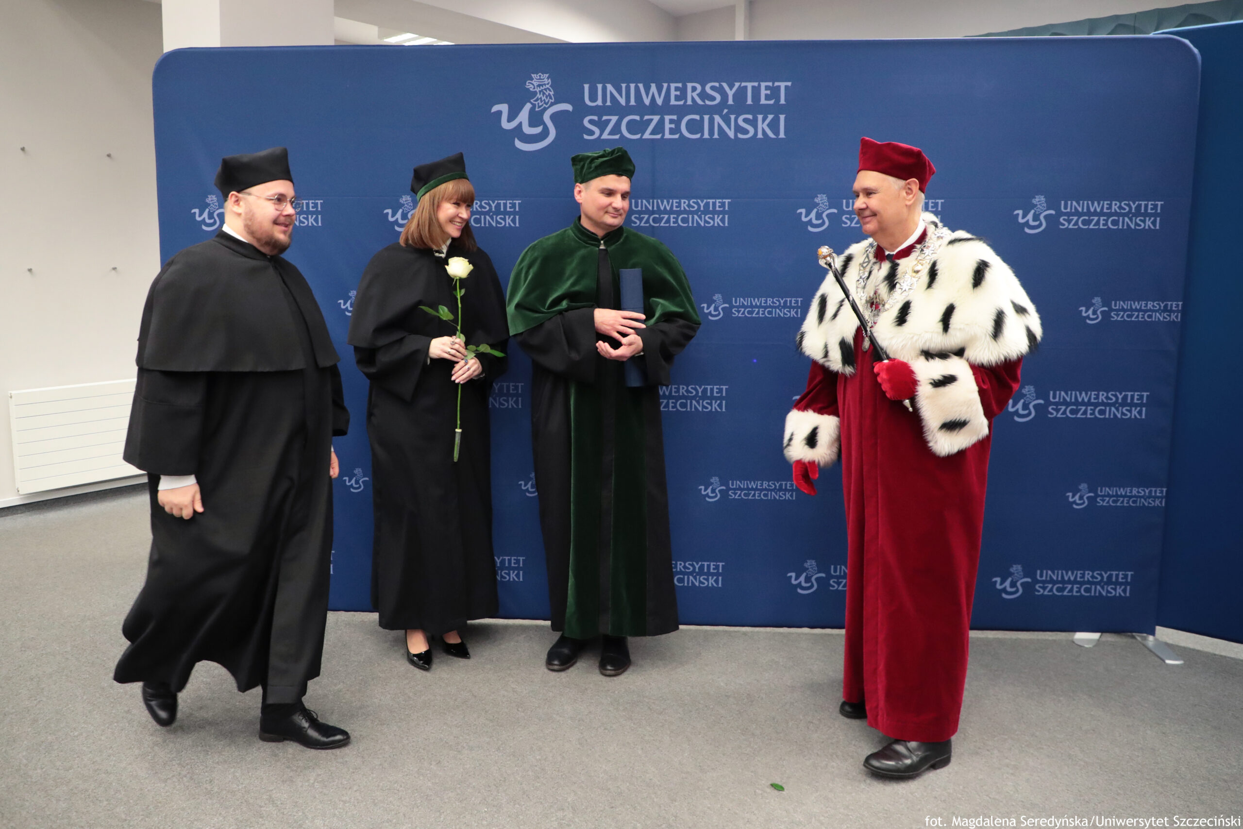 Czworo dorosłych w strojach akademickich stojących przed niebieską ścianką z napisem „UNIWERSYTET SZCZECIŃSKI” i logo uczelni. Po lewej stoi mężczyzna w czarnej todze i czarnym birecie; ma okulary i zarost, jest uśmiechnięty. Obok stoi kobieta w czarnej todze i birecie; trzyma w dłoni białą różę, ma włosy do ramion i uśmiecha się. W środku stoi mężczyzna w ciemnozielonej todze i zielonym nakryciu głowy; trzyma zwinięty, niebieski dyplom lub tubę. Po prawej stoi mężczyzna w bordowej todze i bordowym nakryciu głowy; ma białe futrzane obszycie w czarne plamki oraz bordowe rękawiczki i trzyma laskę lub berło. Na dole po prawej widać podpis fotograficzny: „fot. Magdalena Seredyńska/Uniwersytet Szczeciński”.