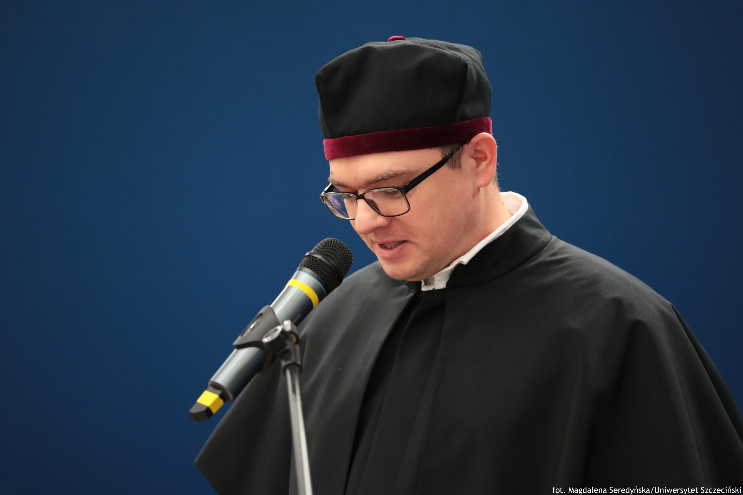 Mężczyzna w czarnej todze i czarnym birecie z bordowym paskiem, stojący przy mikrofonie. Ma jasną skórę, krótkie włosy i okulary w ciemnych oprawkach; głowa jest lekko pochylona, jak podczas czytania lub przemawiania. Mikrofon jest na statywie i ma żółte oznaczenia na obudowie. Tło jest jednolite, granatowe. W prawym dolnym rogu widoczny jest podpis: „fot. Magdalena Seredyńska/Uniwersytet Szczeciński”.