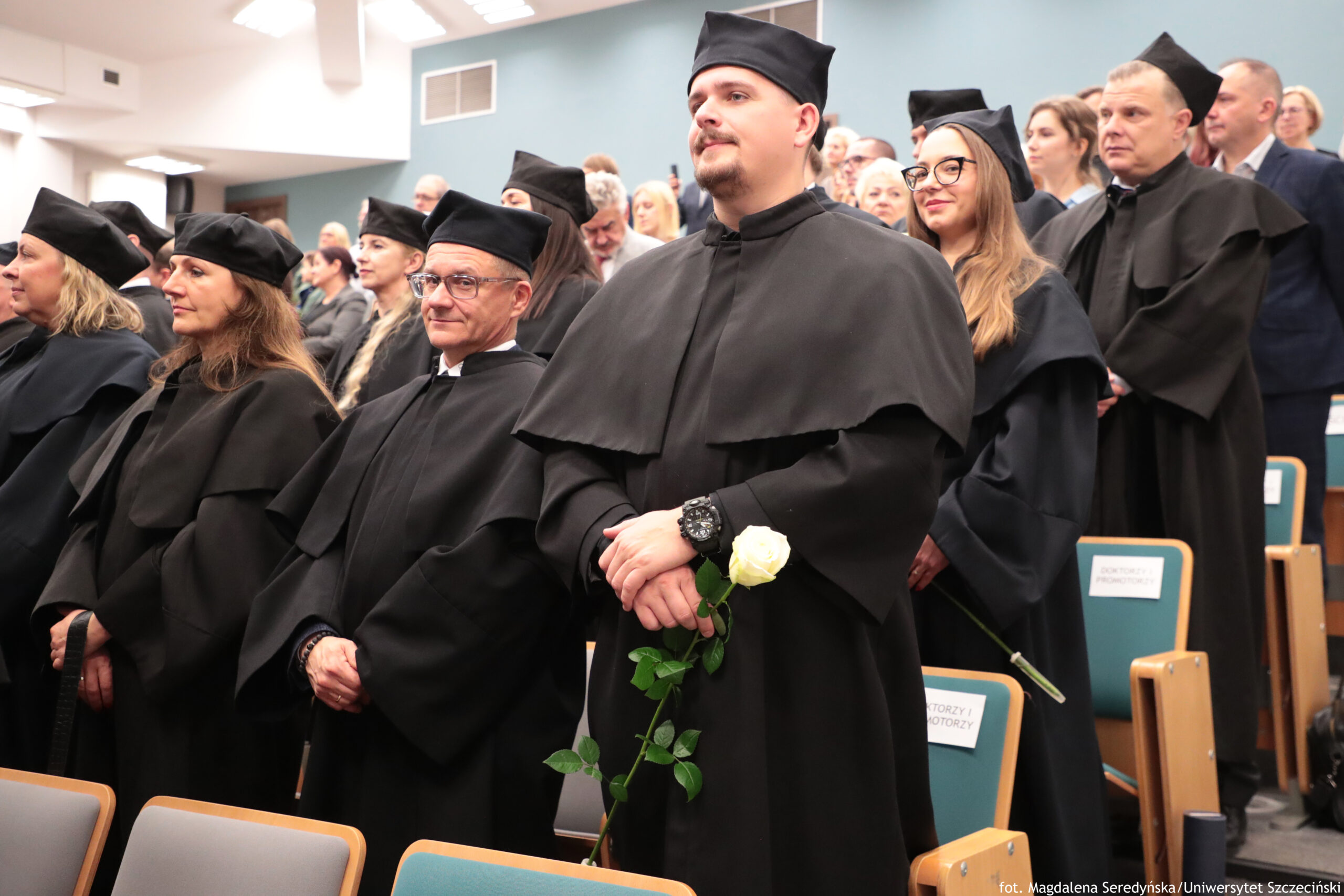 Grupa dorosłych osób w czarnych togach i czarnych biretach stojąca w auli. W pierwszym planie stoi wysoki mężczyzna z krótką brodą, w todze, trzymający białą różę; na nadgarstku ma duży czarny zegarek. Obok i za nim stoją inne osoby w podobnych strojach, w tym kobieta w okularach z długimi włosami. W tle widać kolejne osoby na sali oraz jasnoniebieską ścianę. Na oparciach niektórych krzeseł są kartki z napisem „DOKTORZY I DOKTORZY”. W prawym dolnym rogu jest podpis: „fot. Magdalena Seredyńska / Uniwersytet Szczeciński”.