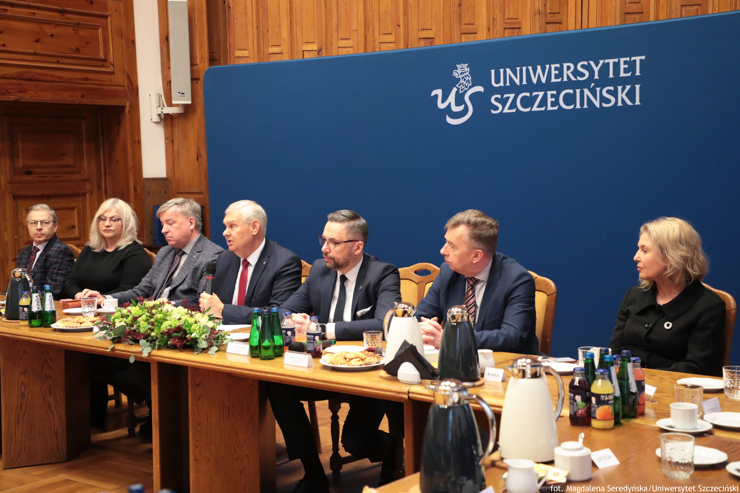 Długi stół konferencyjny z ośmioma osobami siedzącymi obok siebie, przed granatowym tłem z napisem „UNIWERSYTET SZCZECIŃSKI” i logo „US”. Osoby są ubrane formalnie, w garnitury i eleganckie stroje. W centrum siedzi mężczyzna w garniturze z czerwonym krawatem, trzymający mikrofon i wyglądający na przemawiającego; obok niego siedzą inni uczestnicy, część zwrócona w jego stronę. Na stole stoją karafki i termosy do kawy lub herbaty (czarne i jasne), szklanki, filiżanki, talerzyki z ciastkami lub przekąskami, zielone i kolorowe butelki z napojami, oraz małe wizytówki z nazwiskami (tekst na nich jest zbyt mały, by był czytelny). Pośrodku stołu leży duża kompozycja kwiatowa w kolorach zieleni, bieli i czerwieni.