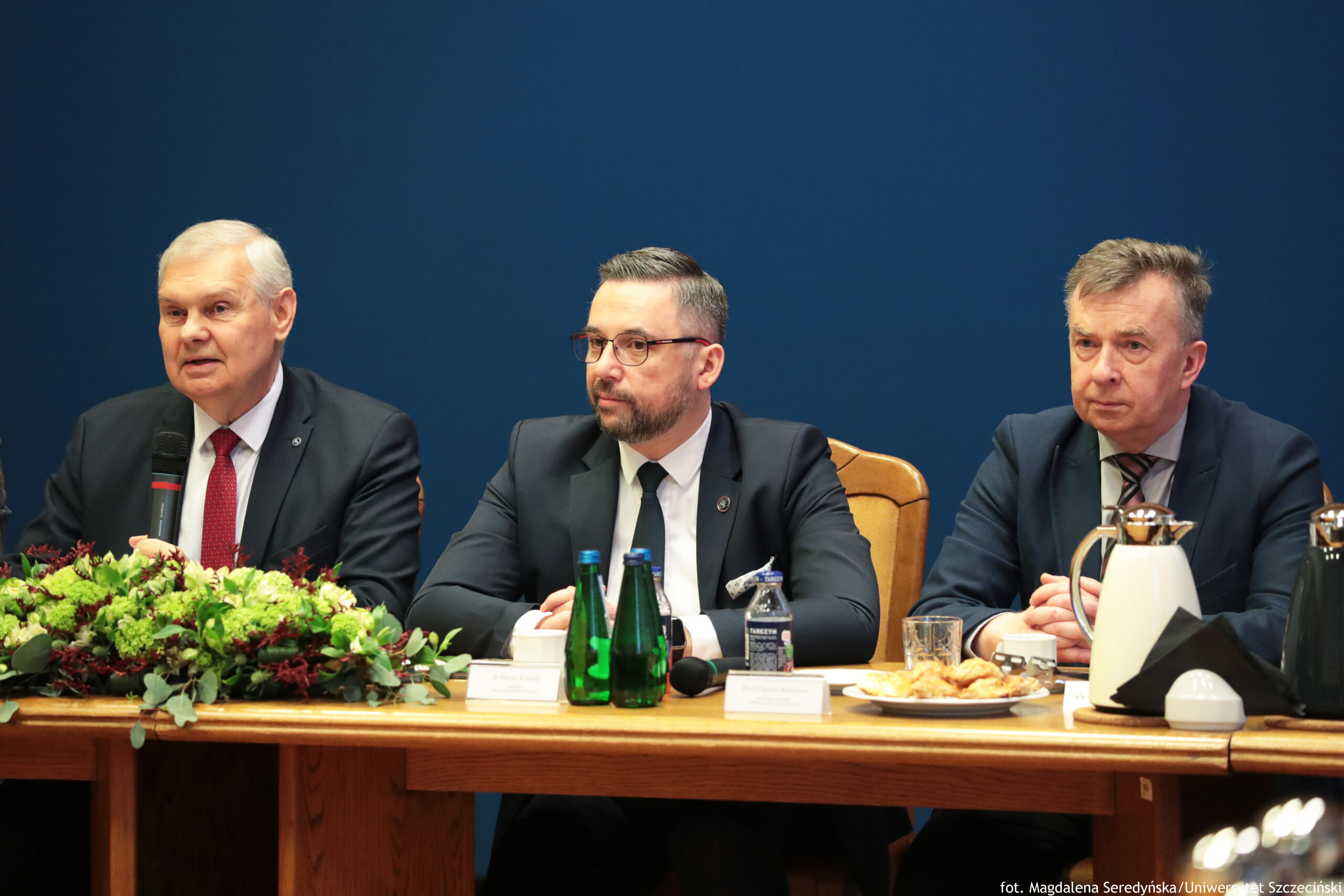 Trzech mężczyzn w garniturach siedzących za długim stołem podczas oficjalnego spotkania. Tło stanowi gładka, ciemnoniebieska ściana. Po lewej stronie siedzi starszy mężczyzna o jasnej skórze i białych włosach, w ciemnym garniturze, białej koszuli i czerwonym krawacie. Trzyma w dłoni mikrofon i wygląda na osobę przemawiającą. Na środku siedzi mężczyzna o jasnej skórze, krótkich ciemnych włosach z siwizną, w okularach, z krótką brodą. Ma na sobie ciemny garnitur, białą koszulę i ciemny krawat; przed nim stoją dwie zielone butelki wody, leży też mikrofon i widać małą butelkę lub kartonik napoju. Przy jego miejscu jest także stojąca tabliczka z nazwiskiem, ale tekst jest nieczytelny. Po prawej stronie siedzi mężczyzna o jasnej skórze i krótkich włosach z siwizną, w ciemnym garniturze, białej koszuli i krawacie w paski. Ma dłonie złożone na stole; przed nim stoi dzbanek/termos i szklanka, a także talerz z drobnymi przekąskami. Na stole, po lewej stronie kadru, znajduje się duża kompozycja roślinna z zielonymi i bordowymi elementami. W prawym dolnym rogu widać mały napis kredytowy: „fot. Magdalena Seredyńska/Uniwersytet Szczeciński”.