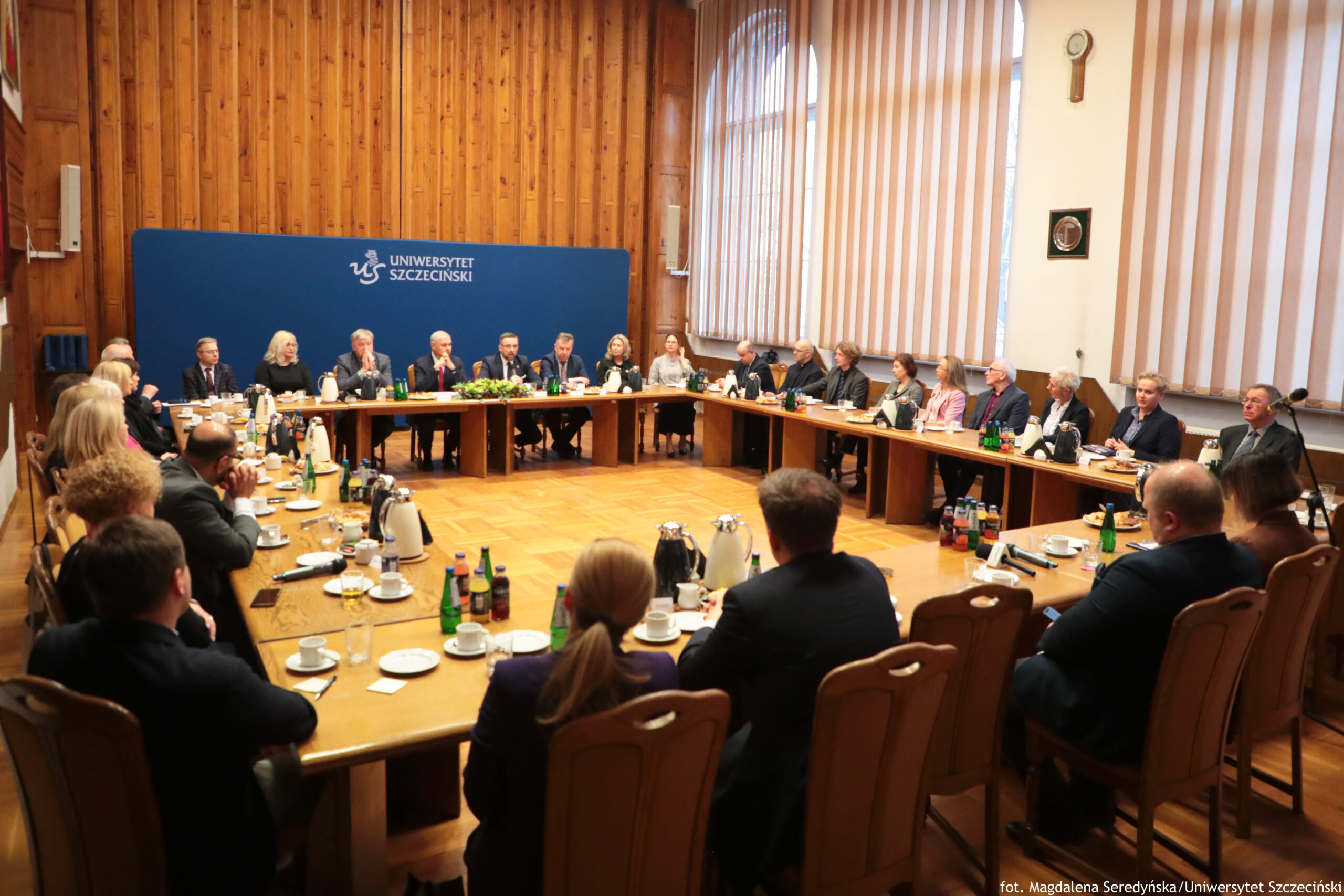 Sala Senatu US z drewnianą boazerią na ścianach i parkietem. W centrum stoi zestaw długich stołów, a wokół nich siedzi kilkadziesiąt osób w strojach formalnych (garnitury, marynarki). Na stole stoją dzbanki termiczne, filiżanki ze spodkami, szklanki, butelki z napojami, talerzyki z przekąskami oraz mikrofony; na środku podkowy widać też dekorację z zieleni. Na tylnej ścianie jest duży niebieski panel z białym napisem i logo: “UNIWERSYTET SZCZECIŃSKI”. Po prawej stronie znajdują się wysokie okna z pionowymi żaluzjami, a na ścianie obok okien wisi okrągły zegar i mała tablica lub emblemat. W prawym dolnym rogu zdjęcia jest podpis: “fot. Magdalena Seredyńska/Uniwersytet Szczeciński”.