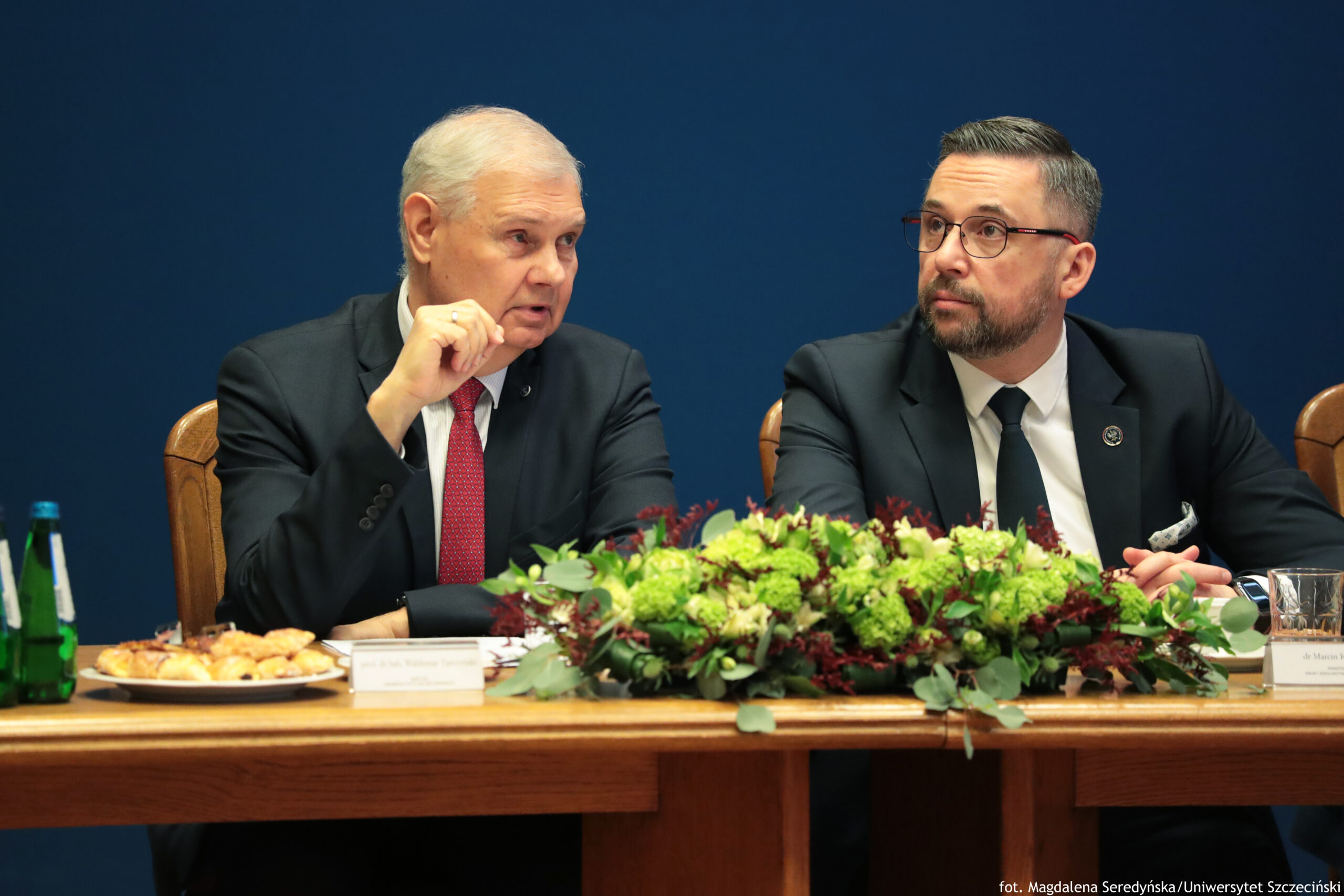 Dwóch mężczyzn w garniturach siedzących przy długim stole konferencyjnym na tle jednolitej, granatowej ściany. Po lewej stronie siedzi starszy mężczyzna o jasnej skórze i siwych włosach, w ciemnym garniturze, białej koszuli i czerwonym krawacie. Ma uniesioną dłoń w geście jak podczas mówienia i jest zwrócony w stronę mężczyzny obok. Po prawej stronie siedzi młodszy mężczyzna o jasnej skórze, krótkich ciemnych włosach, w okularach i z krótką brodą. Ma na sobie ciemny garnitur, białą koszulę i czarny krawat; na klapie marynarki widać okrągłą przypinkę. Patrzy w lewo, w stronę starszego mężczyzny. Na stole znajduje się duża kompozycja roślinna z zielonych i bordowych kwiatów oraz liści. Po lewej widać zieloną butelkę wody, talerz z małymi przekąskami oraz wizytówkę z niewyraźnym tekstem. Po prawej stoi szklanka i kolejna wizytówka. W prawym dolnym rogu jest mały napis: „fot. Magdalena Seredyńska/Uniwersytet Szczeciński”.