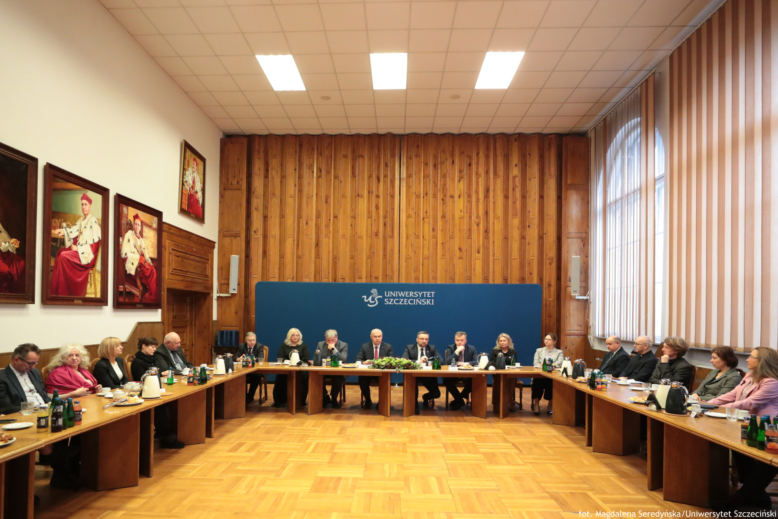 Sala Senatu US z długimi stołami i grupą osób siedzących wokół. W centralnej części, na tle granatowej ścianki z białym napisem „UNIWERSYTET SZCZECIŃSKI” i logo „US”, siedzi kilku uczestników w garniturach i eleganckich strojach, zwróconych w stronę środka stołów. Ściana za nimi jest wyłożona pionowymi, drewnianymi panelami. Po prawej stronie znajdują się wysokie okna z pionowymi żaluzjami; światło dzienne wpada do wnętrza. Na suficie są prostokątne panele oświetleniowe. Podłoga jest drewniana, ułożona w parkiet. Na stołach stoją butelki wody i napojów, filiżanki, dzbanki/termosy, talerzyki z przekąskami oraz dokumenty. Na lewnej ścianie wiszą duże portrety w ozdobnych ramach (osoby w czerwono-białych strojach). W prawym dolnym rogu zdjęcia widoczny jest podpis: „fot. Magdalena Seredyńska / Uniwersytet Szczeciński”.