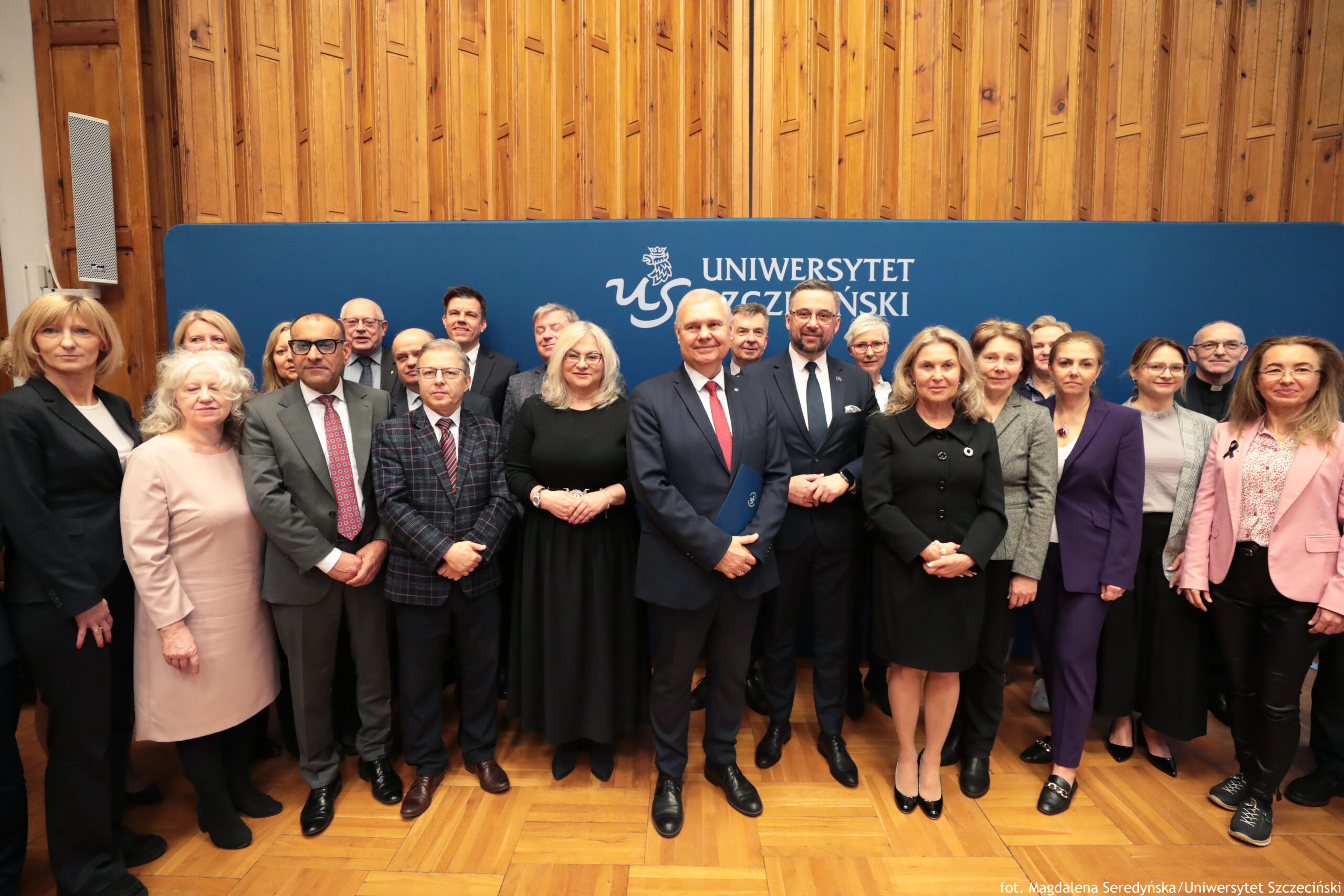 Grupa kilkunastu osób pozujących do wspólnego zdjęcia na tle granatowej ścianki z napisem „UNIWERSYTET SZCZECIŃSKI” i białym logo z gryfem. Osoby stoją w dwóch rzędach, większość w strojach formalnych: mężczyźni w garniturach i krawatach, kobiety w garsonkach, marynarkach lub eleganckich sukienkach. W pierwszym rzędzie, mniej więcej na środku, stoją dwaj mężczyźni w ciemnych garniturach; jeden ma czerwony krawat i trzyma niebieską teczkę, drugi ma czarny krawat i okulary. Obok nich stoją kobiety w ciemnych strojach, w tym jedna w czarnej sukience oraz jedna w ciemnym płaszczu/sukience. Po lewej stronie pierwszego rzędu widać m.in. kobietę w jasnoróżowej sukience oraz mężczyznę w szarym garniturze z czerwonym krawatem i okularami. Po prawej stronie stoją m.in. kobieta w fioletowym garniturze oraz kobieta w różowej marynarce i czarnych spodniach. Tło to drewniana ściana z pionowymi panelami i parkietowa podłoga. W prawym dolnym rogu jest podpis: „fot. Magdalena Seredyńska/Uniwersytet Szczeciński”.
