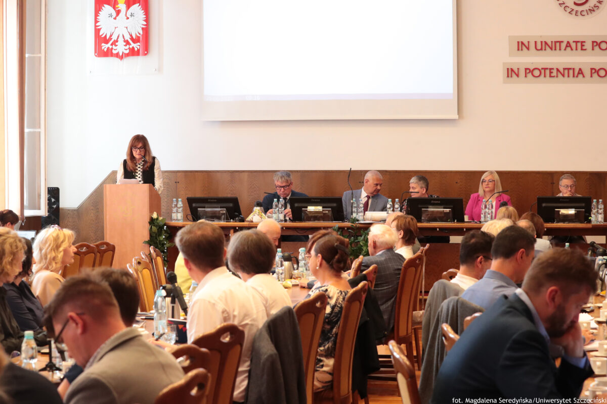 Sala Senatu US z długimi stołami, krzesłami i dużą liczbą uczestników siedzących tyłem lub bokiem do kamery. W tle, przy mównicy, stoi przemawiająca kobieta z długimi włosami i okularami. Za nią, przy długim stole prezydialnym, siedzi kilka osób (kobiety i mężczyźni), przed którymi stoją monitory, mikrofony i butelki wody. Na ścianie nad mównicą wisi godło Polski (biały orzeł na czerwonym tle), a wyżej znajduje się duży ekran projekcyjny. Po prawej stronie ściany widoczny jest fragment napisu po łacinie, zaczynający się od „IN UNITATE …” oraz „IN POTENTIA …”. Na dole po prawej widnieje podpis fotografa: „fot. Magdalena Seredyńska/Uniwersytet Szczeciński”.