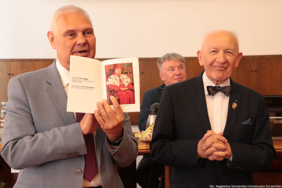 Dwóch starszych mężczyzn w garniturach podczas posiedzenia Senatu US. Po lewej mężczyzna w jasnoszarym garniturze i bordowym krawacie trzyma otwartą książkę i pokazuje jej stronę z tekstem oraz kolorową ilustracją osoby w czerwonym stroju (odświętnym stroju Rektora US). Po prawej mężczyzna w czarnym garniturze, białej koszuli i ciemnej muszce w kropki stoi z dłońmi złożonymi z przodu; ma przypiętą odznakę w klapie. W tle widać siedzącego mężczyznę oraz drewniane elementy sali. Widoczny tekst w książce (lewa strona): POCZET REKTORÓW (1986–2025) UNIWERSYTETU SZCZECIŃSKIEGO DZIEJE DR. NAUK MEDYCZNYCH MIECZYSŁAWA CHRUŚCIELA Widoczny tekst w prawym dolnym rogu zdjęcia: fot. Magdalena Seredyńska/ Uniwersytet Szczeciński