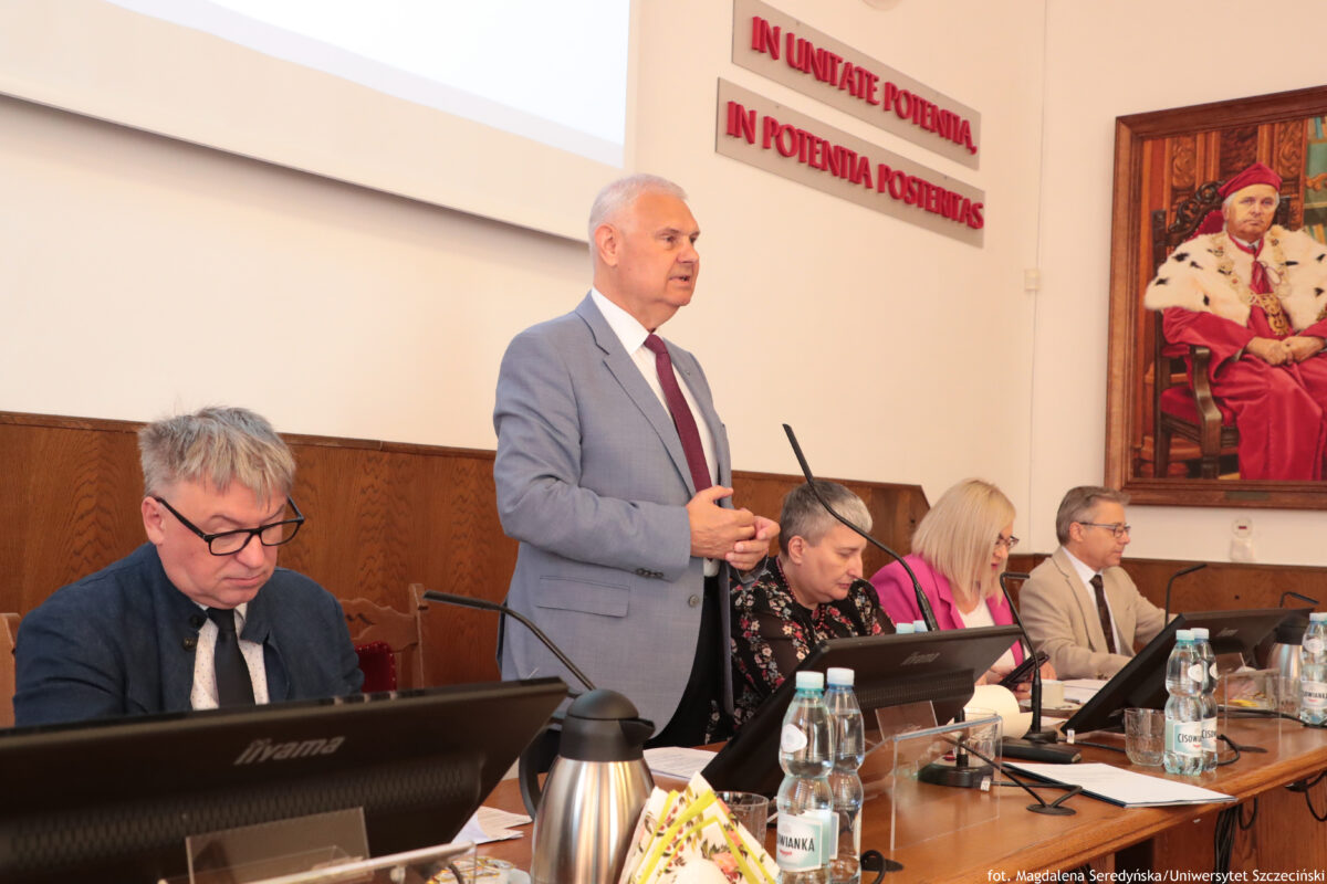 Sala Senatu US z długim stołem prezydialnym i kilkoma osobami przy mikrofonach oraz monitorach. Na środku za stołem stoi starszy mężczyzna z krótkimi siwymi włosami, w jasnoszarym garniturze i bordowym krawacie; wygląda na przemawiającego. Po lewej siedzi mężczyzna w okularach, z siwymi włosami, w ciemnej marynarce, pochylony nad dokumentami. Po prawej stronie przy stole siedzą trzy osoby: kobieta z siwymi krótkimi włosami w ciemnym ubraniu, kobieta z blond włosami i okularami w jasnym ubraniu oraz mężczyzna w jasnej marynarce i okularach. Na stole widać monitory, mikrofony na długich wysięgnikach, butelki wody, szklanki, dzbanek/termos i rozłożone papiery. Na ścianie w tle są dwa napisy z czerwonych liter: „IN UNITATE POTENTIA” oraz „IN POTENTIA POSTERITAS”. Po prawej wisi duży portret osoby w czerwonym stroju z białym futrzanym obszyciem. W prawym dolnym rogu jest podpis: „fot. Magdalena Seredyńska / Uniwersytet Szczeciński”.