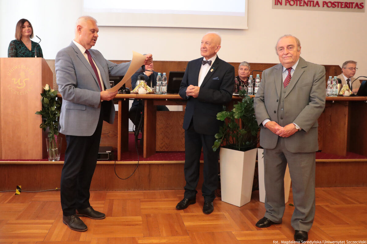 Uroczystość w sali Senatu US z trzema starszymi mężczyznami w garniturach na pierwszym planie oraz stołem prezydialnym w tle. Po lewej stoi mężczyzna w jasnoszarym garniturze z bordowym krawatem, czyta z kartki trzymanej w rękach. Na środku stoi łysy mężczyzna w ciemnym garniturze z białą koszulą i czarną muchą, ma ręce złożone przed sobą i jest zwrócony w stronę mężczyzny czytającego. Po prawej stoi mężczyzna w szarym trzyczęściowym garniturze z czerwonym krawatem w szare paski, ma ręce splecione przed sobą i patrzy w stronę aparatu. W tle widać długi stół z siedzącymi osobami, butelkami wody i papierami. Z lewej strony jest mównica z logo uczelni i mikrofonem; za nią stoi kobieta w ciemnej sukience we wzory. Na podłodze przy stole stoją białe donice z zielonymi roślinami. W prawym dolnym rogu jest podpis: „fot. Magdalena Seredyńska / Uniwersytet Szczeciński”.