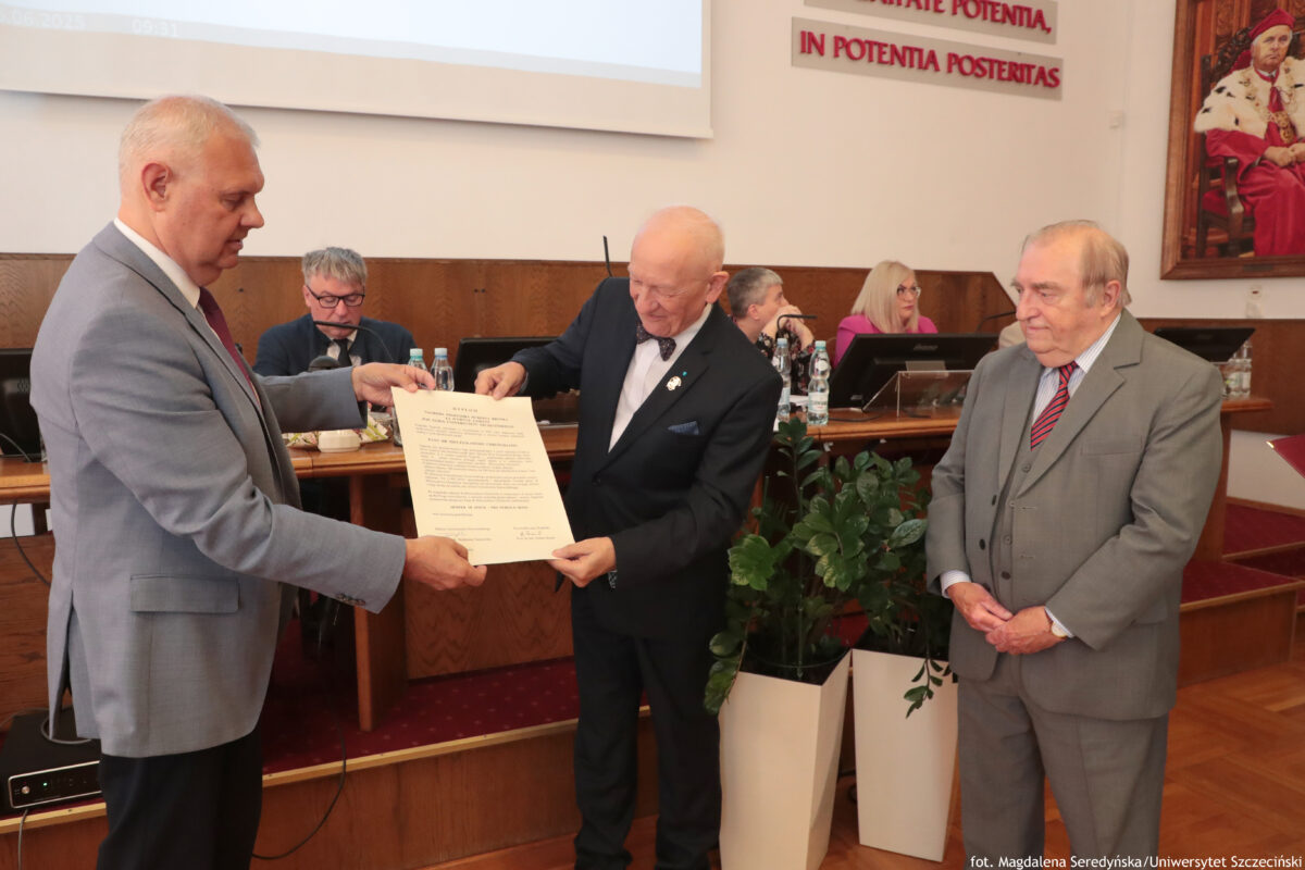 Na zdjęciu w sali senatu US trzech elegancko ubranych mężczyzn przed stołem prezydialnym. Dwóch z nich trzyma w rękach oficjalny dokument – starszy mężczyzna w ciemnym garniturze wręcza go mężczyźnie w jasnoszarym garniturze. Obok stoi trzeci mężczyzna w szarym trzyczęściowym garniturze i czerwonym krawacie, obserwując uroczystość. W tle widać osoby siedzące przy stole prezydialnym z mikrofonami i komputerami oraz dużą roślinę doniczkową. Na ścianie znajduje się napis w języku łacińskim oraz portret w czerwonej todze.