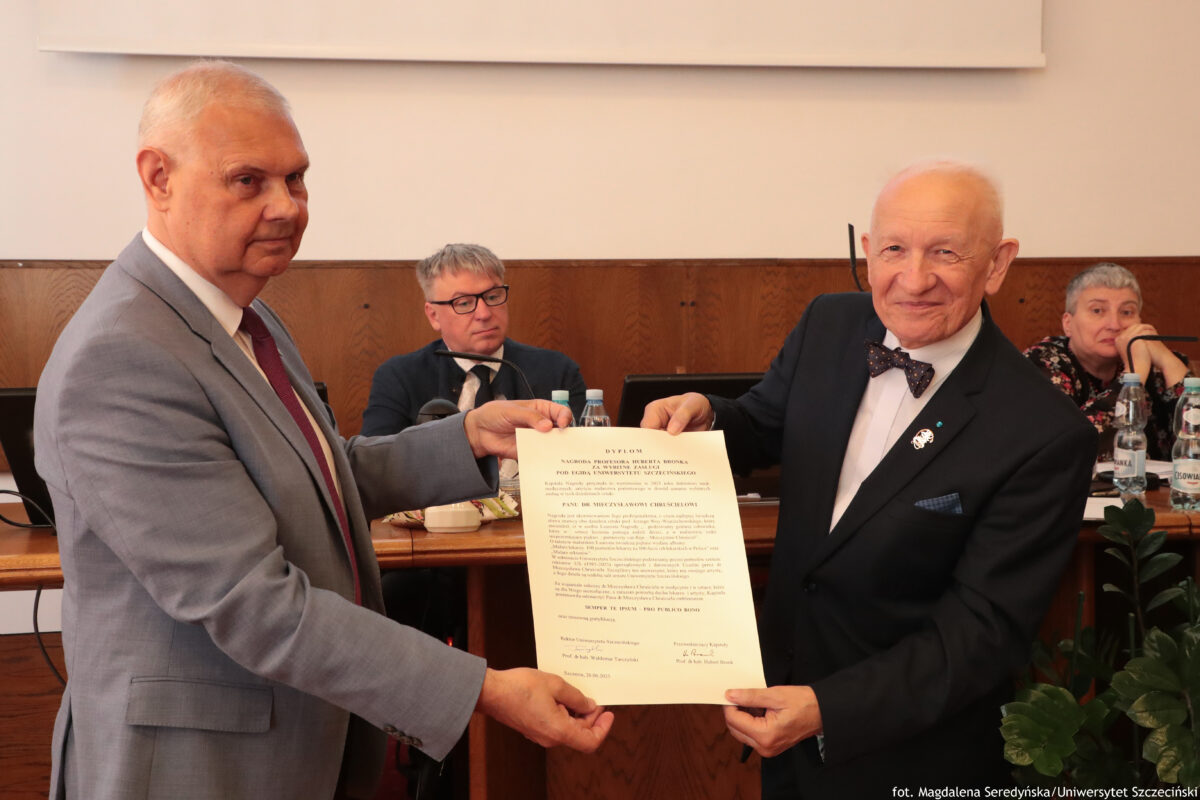 Na zdjęciu dwóch mężczyzn stoi naprzeciw siebie w sali konferencyjnej i wspólnie trzyma oficjalny dokument skierowany w stronę obiektywu. Mężczyzna po lewej ma na sobie jasnoszary garnitur i bordowy krawat, a po prawej – ciemny garnitur z muszką i przypinką w klapie. W tle przy stole siedzą inne osoby z mikrofonami, a za nimi widać drewnianą ścianę oraz fragment ekranu projekcyjnego.