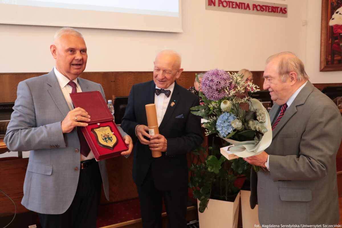 Na zdjęciu w sali senatu US trzech elegancko ubranych mężczyzn stoi obok siebie podczas uroczystości. Mężczyzna po lewej w jasnoszarym garniturze prezentuje otwarte czerwone etui z pamiątkową plakietą. Mężczyzna w środku, ubrany w ciemny garnitur z muszką, trzyma zwinięty dokument w tubie. Po prawej stronie trzeci mężczyzna w szarym garniturze i czerwonym krawacie trzyma duży bukiet kwiatów w jasnym papierze. W tle widać stół prezydialny, mikrofony oraz fragment napisu na ścianie.