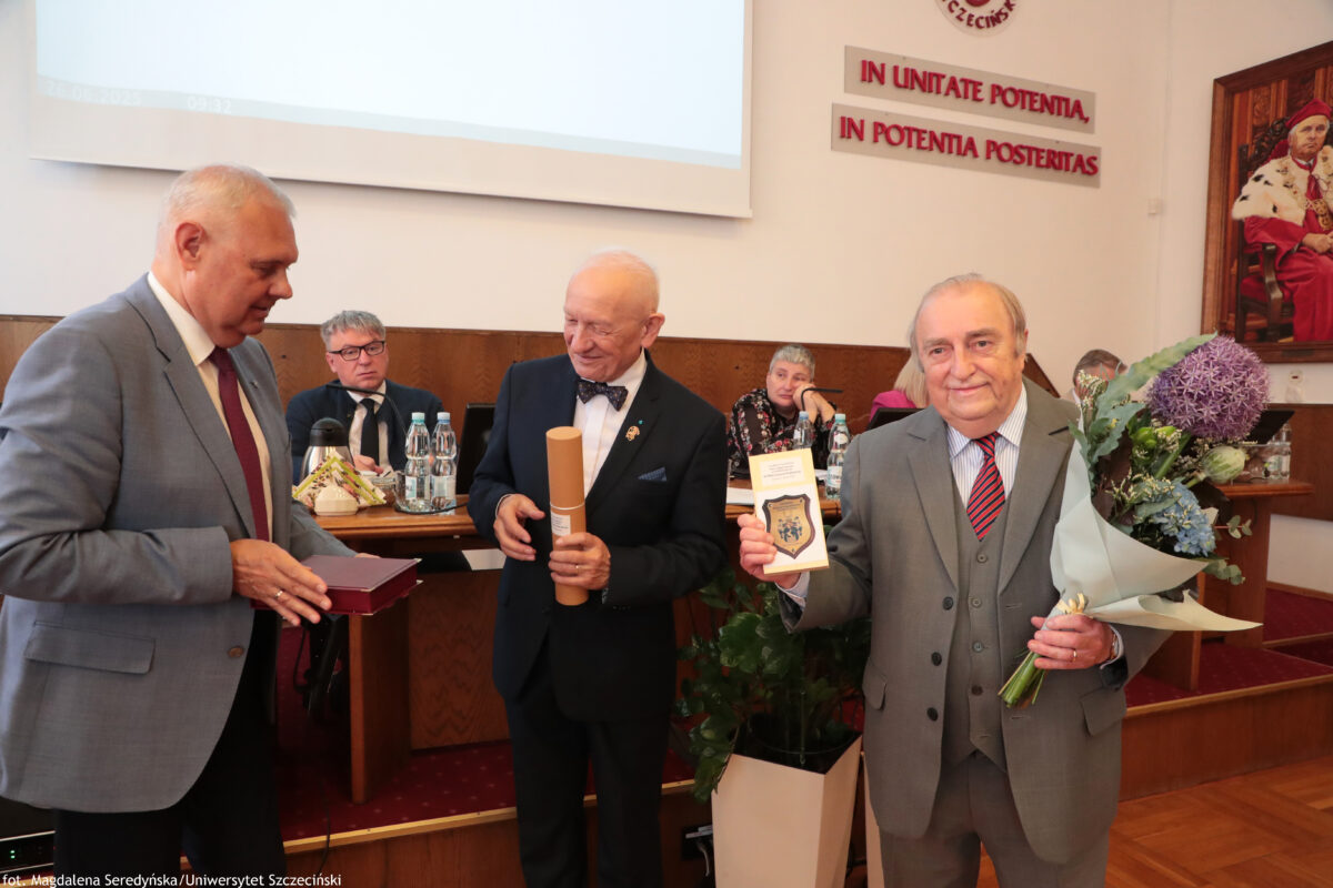 Na zdjęciu w sali senatu US trzech elegancko ubranych mężczyzn stoi obok siebie podczas uroczystości. Mężczyzna po lewej w jasnoszarym garniturze trzyma czerwone pudełko. Mężczyzna w środku, ubrany w ciemny garnitur z muszką, trzyma zwinięty dokument w tubie. Po prawej stronie trzeci mężczyzna w szarym garniturze i czerwonym krawacie trzyma duży bukiet kwiatów w jasnym papierze. W tle widać stół prezydialny, mikrofony oraz fragment napisu na ścianie.