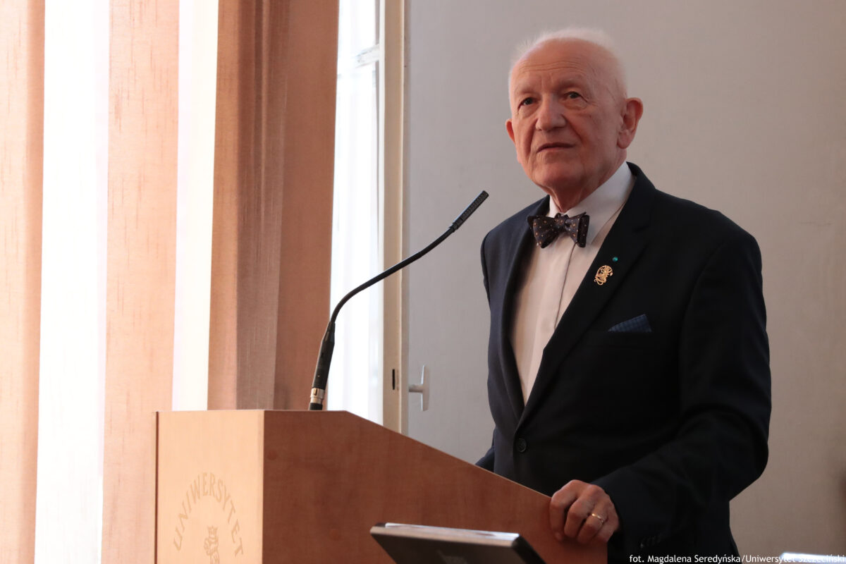 Starszy mężczyzna o jasnej karnacji i białych, krótkich włosach stoi przy drewnianej mównicy z mikrofonem. Ma na sobie ciemny garnitur, białą koszulę i ciemną muchę w drobny wzór. Na lewej klapie marynarki widać małą przypinkę i ozdobę, w kieszonce jest poszetka. Prawa dłoń spoczywa na krawędzi mównicy, na palcu jest obrączka. Tło stanowi jasna ściana i wysokie okno z beżową zasłoną, przez które wpada światło dzienne. Na froncie mównicy widać słabo czytelny napis „UNIWERSYTET”. W prawym dolnym rogu jest podpis: „fot. Magdalena Seredyńska/Uniwersytet Szczeciński”.