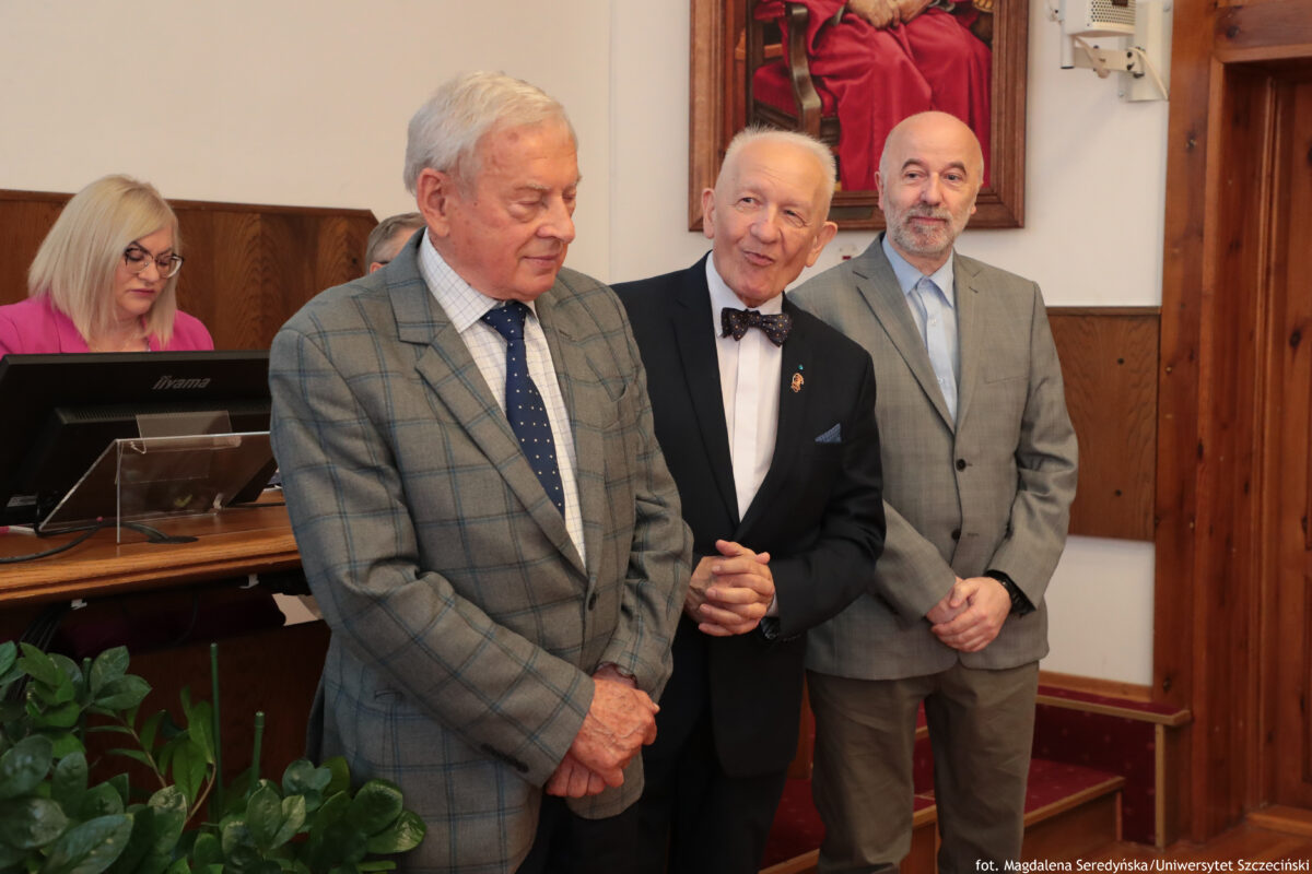 Na pierwszym planie po lewej stoi starszy mężczyzna o jasnej skórze i siwych włosach, w szarym garniturze w kratę, białej koszuli i granatowym krawacie w drobne kropki. Ma ręce złożone z przodu i patrzy w dół. Na środku stoi starszy mężczyzna o jasnej skórze i krótkich siwych włosach, w czarnym garniturze, białej koszuli i ciemnej muszce w kropki. Ma przypiętą małą odznakę na klapie marynarki i uśmiecha się, zwrócony w stronę mężczyzny po lewej. Po prawej stoi mężczyzna o jasnej skórze, łysy, z krótką siwą brodą, w jasnoszarym garniturze i jasnej koszuli. Ma ręce splecione z przodu i lekko się uśmiecha. W tle po lewej siedzi kobieta o jasnej skórze i jasnych blond włosach, w okularach i różowej marynarce, przy stole prezydialnym z monitorem. Na ścianie z tyłu wisi obraz z postacią w czerwonym stroju. W prawym dolnym rogu jest podpis: „fot. Magdalena Seredyńska/Uniwersytet Szczeciński”.
