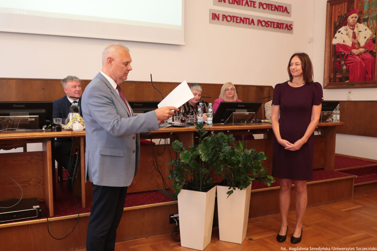 Sala Senatu US z długim stołem prezydialnym i kilkoma osobami siedzącymi za monitorami. Mężczyzna w jasnoszarym garniturze stoi po lewej stronie i czyta z kartki. Po prawej stronie stoi kobieta w bordowej, krótkorękawowej sukience, z rękami złożonymi z przodu, uśmiechnięta. Na pierwszym planie stoją dwie wysokie białe donice z zielonymi roślinami. Na ścianie u góry widoczny jest czerwony napis: „IN UNITATE POTENTIA, IN POTENTIA POSTERITAS”. Po prawej wisi obraz przedstawiający mężczyznę w czerwonym stroju ceremonialnym z białym futrzanym kołnierzem. Na dole po prawej jest mały podpis: „fot. Magdalena Seredyńska/Uniwersytet Szczeciński”.