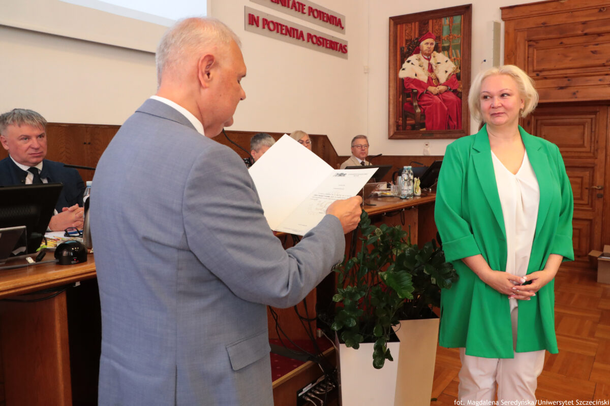 Sala Senatu US z kilkoma osobami przy długich stołach oraz dwiema osobami stojącymi na pierwszym planie. Po lewej stronie stoi mężczyzna w jasnoszarym garniturze, z siwymi, krótko ostrzyżonymi włosami, zwrócony bokiem. Trzyma otwartą teczkę lub dokumenty i czyta wyróżnienie. Po prawej stronie stoi kobieta o jasnej cerze, z krótkimi, jasnoblond włosami. Ma na sobie zieloną marynarkę, białą bluzkę i białe spodnie. Ręce ma złożone z przodu i ma lekki uśmiech. W tle widać kilku mężczyzn w garniturach siedzących przy stołach prezydialnym za monitorami. Na ścianie jest duży obraz przedstawiający mężczyznę w czerwonych szatach, siedzącego na tronie. Nad stołami znajduje się napis na ścianie: „UNITATE POTENTIA, IN POTENTIA POTESTAS”. Na dole po prawej jest podpis fotograficzny: „fot. Magdalena Seredyńska/Uniwersytet Szczeciński”.