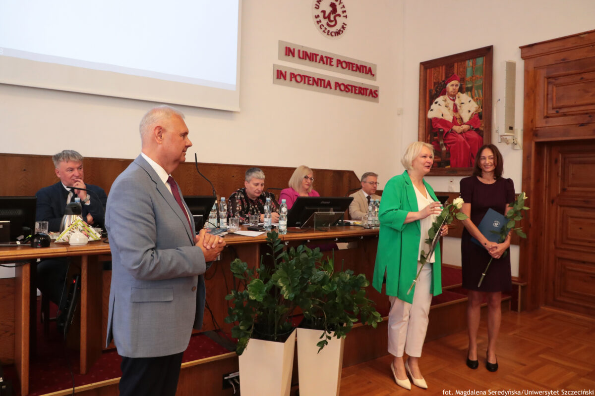 Sala konferencyjna z długim stołem prezydialnym i kilkoma osobami siedzącymi za mikrofonami i monitorami. Na pierwszym planie stoi mężczyzna w jasnoszarym garniturze, zwrócony bokiem, jak podczas przemówienia lub podziękowań. Po prawej stoją dwie kobiety: jedna w zielonej marynarce i białych spodniach trzyma białe kwiaty, druga w ciemnej sukience trzyma bukiet oraz niebieską teczkę. Na ścianie w tle widać łaciński napis „IN UNITATE POTENTIA, IN POTENTIA POSTERITAS” oraz duży portret mężczyzny w czerwonym stroju ceremonialnym. W dolnej części kadru stoją duże donice z zielonymi roślinami. W prawym dolnym rogu jest podpis: „fot. Magdalena Seredyńska/Uniwersytet Szczeciński”.