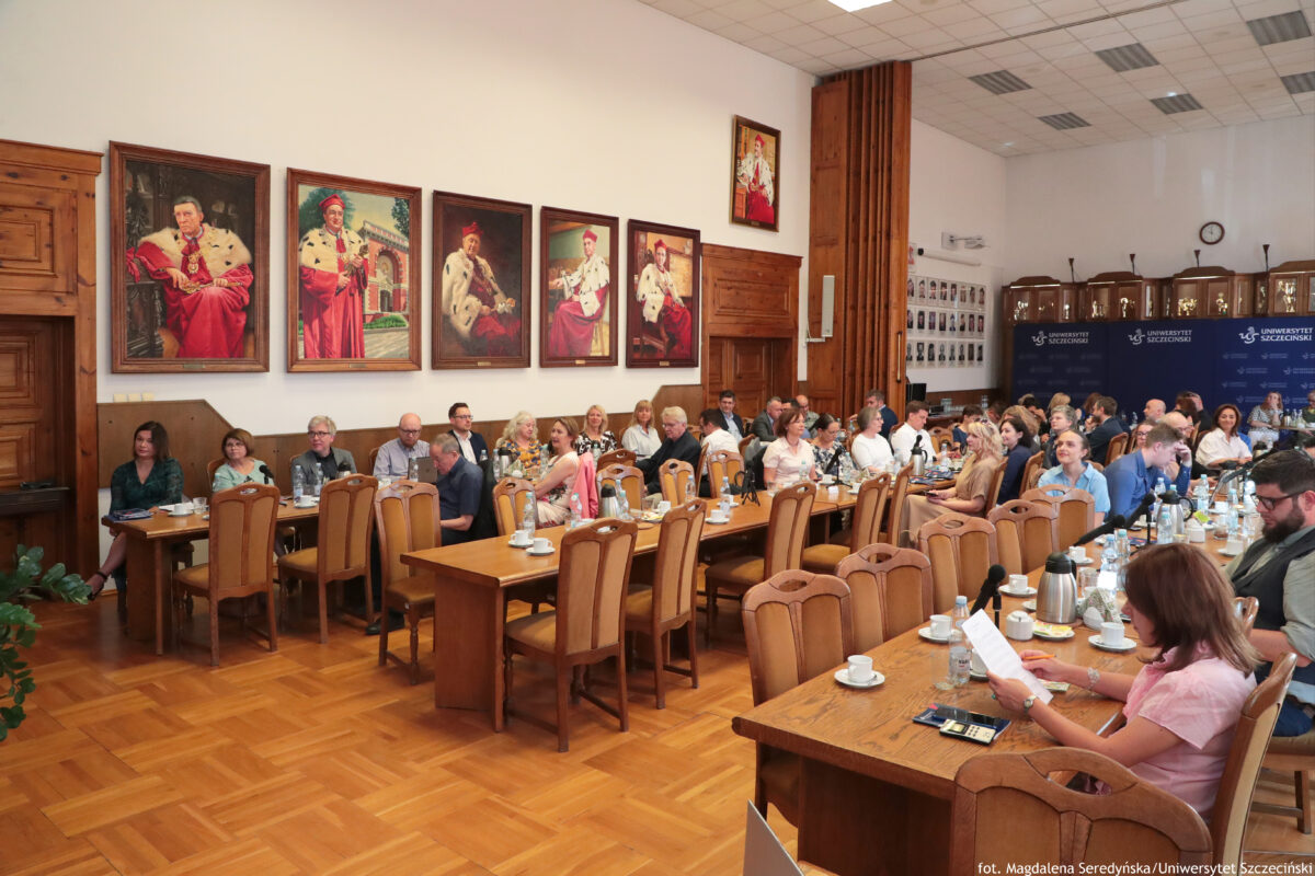 Duża sala Senatu US z długimi drewnianymi stołami ustawionymi w podkowę i wieloma osobami siedzącymi przy nich. Na stołach stoją białe filiżanki ze spodkami, butelki wody, dzbanki/termosy i mikrofony konferencyjne; część osób ma przed sobą kartki i notatki. Na lewej ścianie wisi kilka dużych portretów w ramach, przedstawiających mężczyzn w czerwonych strojach ceremonialnych z białymi gronostajowymi pelerynami. W głębi po prawej stronie stoją granatowe ścianki z napisami „UNIWERSYTET SZCZECIŃSKI” i logo uczelni. Na ścianie obok widać tablicę z wieloma małymi zdjęciami (prawdopodobnie portretami). W prawym dolnym rogu zdjęcia siedzi kobieta w różowej bluzce, trzymająca kartkę. Widoczny podpis na dole: „fot. Magdalena Seredyńska/Uniwersytet Szczeciński”.