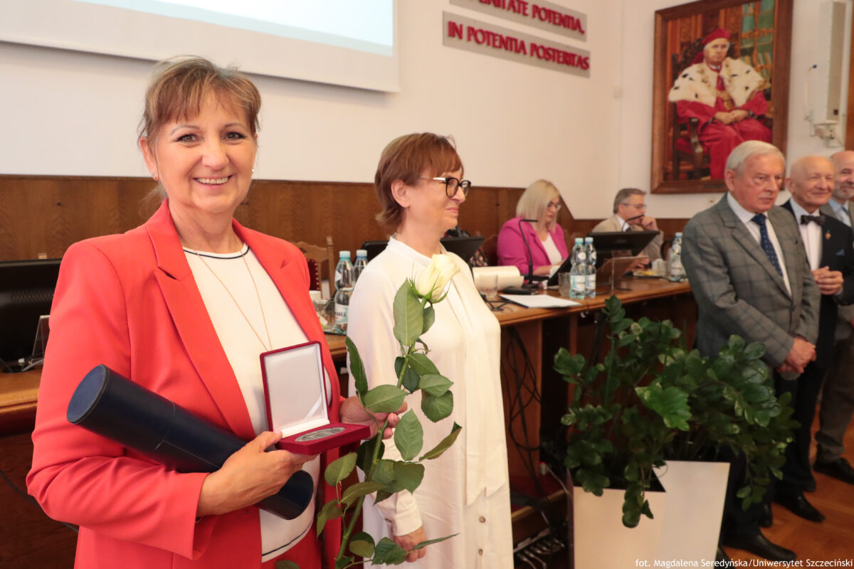 Kobieta w koralowym żakiecie uśmiechająca się do aparatu, trzymająca białą różę oraz otwarte czerwone pudełko z medalem, a także granatową tubę. Ma upięte brązowe włosy i jasną karnację, ubrana jest też w białą bluzkę i cienki naszyjnik. Scena wygląda na uroczystość w sali Senatu US: w tle stoją i siedzą inne osoby w strojach formalnych, na długim stole są butelki z wodą i monitory. Na ścianie wisi duży portret mężczyzny w czerwonym, ceremonialnym stroju. Nad stołem widoczny jest napis po łacinie: “IN POTENTIA POSSE NOSTRAS”. W prawym dolnym rogu jest podpis: “fot. Magdalena Seredyńska / Uniwersytet Szczeciński”.
