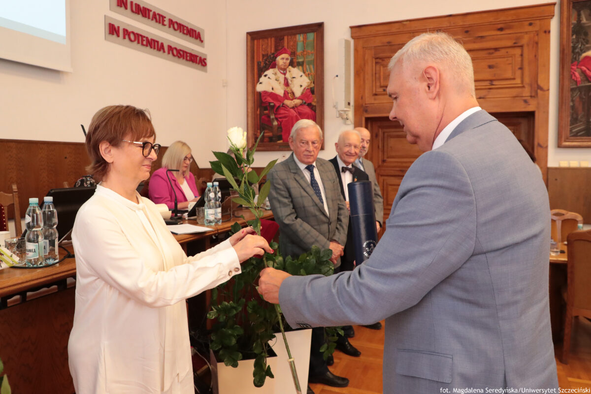 Uroczystość w sali Senatu US, z kilkoma osobami w tle i obrazami na ścianach. Na pierwszym planie po lewej stronie stoi kobieta w białej bluzce, w okularach, z krótkimi brązowymi włosami. Po prawej stronie stoi mężczyzna w jasnoszarym garniturze, z krótkimi siwymi włosami. Między nimi przekazywana jest pojedyncza biała róża z długą łodygą. W tle stoji trzech starszych mężczyzn w garniturach, jeden w szarym garniturze w kratę, drugi z muchą, trzeci w szarym garniturze, patrzą w stronę pierwszego planu. Po lewej stronie przy stole siedzi kobieta w różowej marynarce; na stole stoją butelki wody i dokumenty. Na ścianie z tyłu widać łaciński napis: IN UNITATE POTENTIA, IN POTENTIA POSENTAS Na ścianie wiszą też duże obrazy, w tym portret osoby w czerwonym stroju. W prawym dolnym rogu jest podpis: fot. Magdalena Seredyńska/Uniwersytet Szczeciński.