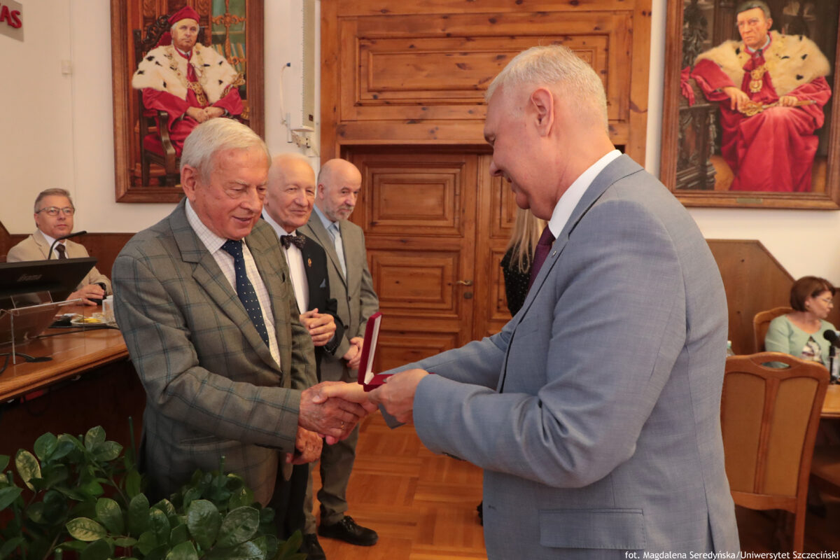 Uroczysta scena wręczenia odznaczenia w sali Senatu US. Na pierwszym planie dwaj starsi mężczyźni o jasnej karnacji ściskają sobie dłonie. Mężczyzna po lewej ma siwe włosy, szarą marynarkę w kratę i granatowy krawat w kropki. Mężczyzna po prawej ma krótkie siwe włosy i jasnoszary garnitur; trzyma otwarte czerwone etui z odznaczeniem lub medalem. W tle stoją dwie osoby w garniturach, a po bokach widać uczestników siedzących przy stołach. Na ścianach wiszą dwa duże portrety mężczyzn w czerwonych, ceremonialnych strojach. Na dole po lewej widać zieloną roślinę doniczkową. W prawym dolnym rogu jest podpis: „fot. Magdalena Seredyńska/Uniwersytet Szczeciński”.