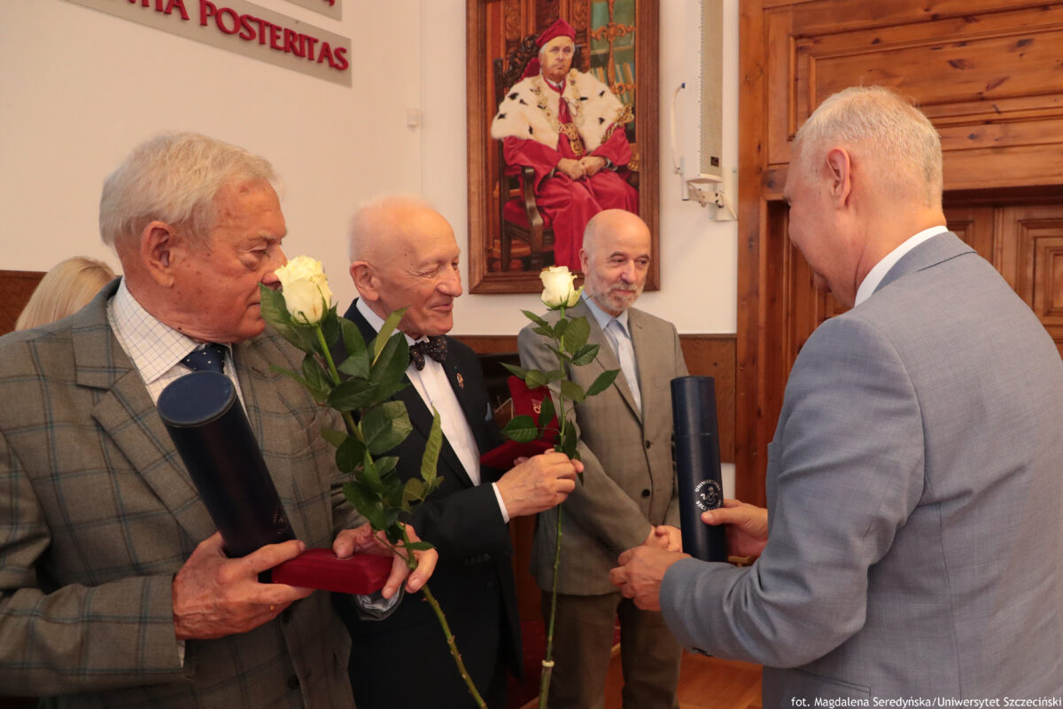 Grupa czterech mężczyzn w eleganckich garniturach w pomieszczeniu, podczas uroczystego wręczania kwiatów i upominków. Po lewej stoi starszy mężczyzna o siwych włosach, w szarym garniturze w kratę; trzyma długą, granatową tubę oraz czerwone etui. Obok niego starszy, łysy mężczyzna w ciemnym garniturze i muszce trzyma białą różę. W tle stoi trzeci mężczyzna, również łysy, z siwą brodą, w jasnoszarym garniturze. Po prawej, odwrócony bokiem do aparatu, stoi mężczyzna w jasnym, niebieskoszarym garniturze; trzyma podobną granatową tubę i składa gratulacje. Na ścianie w tle wisi duży portret męższczyzny w czerwonym stroju, siedzącego na ozdobnym krześle. W górnej części ściany widać fragment napisu „… POSTERITAS”. Po prawej są drewniane drzwi. Na dole po prawej widoczny jest podpis fotograficzny: „fot. Magdalena Seredyńska/Uniwersytet Szczeciński”.