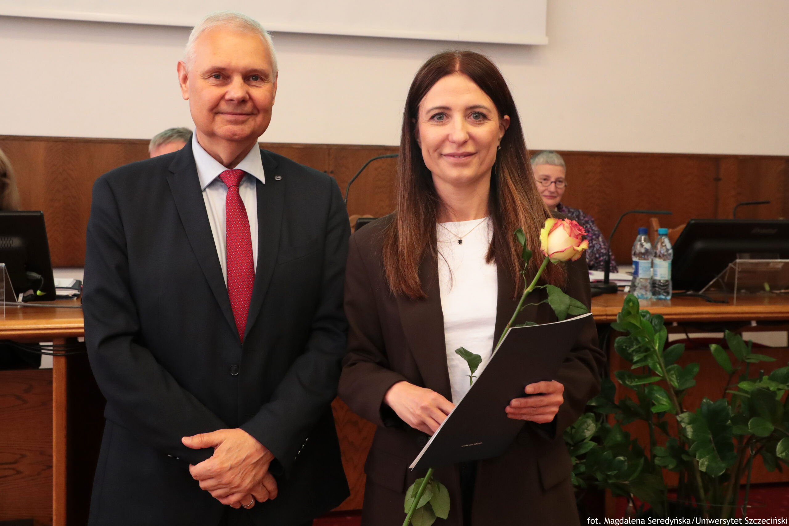 Dwoje dorosłych ludzi stojących obok siebie w sali konferencyjnej. Po lewej mężczyzna o jasnej skórze i siwych włosach, ubrany w ciemny garnitur, jasnoniebieską koszulę i czerwony krawat, patrzy prosto w obiektyw i ma lekki uśmiech. Po prawej kobieta o jasnej skórze i prostych, długich, brązowych włosach, ubrana w ciemnobrązową marynarkę i białą bluzkę, trzyma w rękach pojedynczą różę w odcieniach żółto-różowych oraz ciemną teczkę lub dyplom. W tle widoczny jest drewniany stół konferencyjny z monitorami komputerów, mikrofonami, butelkami wody i dokumentami; za stołem siedzi starsza kobieta z krótkimi siwymi włosami w kolorowej bluzce.