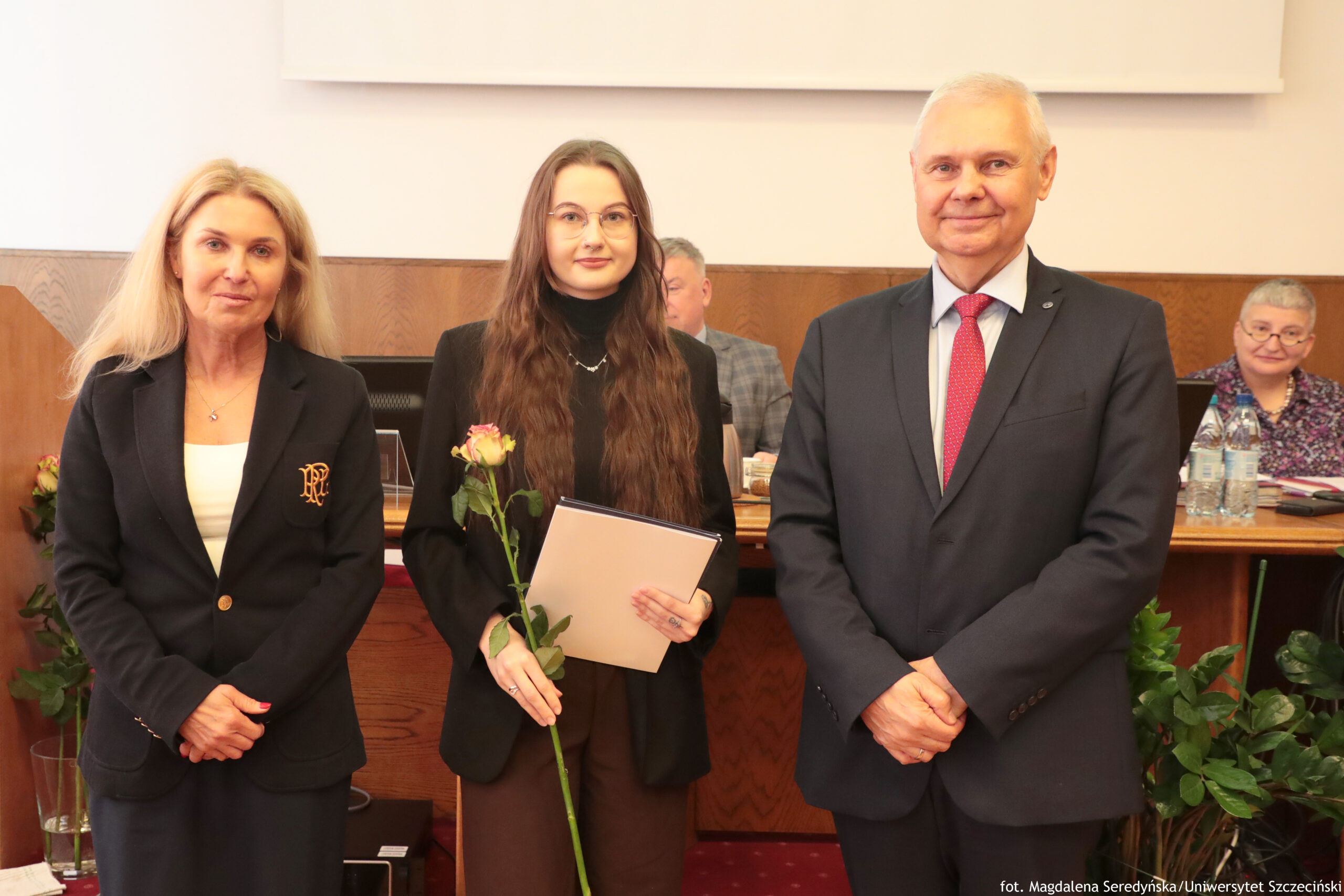 Trzy osoby stojące obok siebie w pomieszczeniu urzędowym lub akademickim. Po lewej stoi kobieta o jasnych, długich włosach, ma jasną skórę i nosi ciemny żakiet z emblematem na lewej piersi oraz jasny top pod spodem. Patrzy w stronę aparatu z neutralnym wyrazem twarzy, dłonie trzyma splecione z przodu. Na środku stoi młoda kobieta o jasnej skórze i bardzo długich, falowanych, brązowych włosach, w okularach. Ubrana jest w czarny golf i ciemną marynarkę, a w rękach trzyma pojedynczą różę o różowo‑kremowym kwiatostanie oraz jasną teczkę lub dyplom. Po prawej stronie stoi starszy mężczyzna o jasnej skórze i krótkich, siwych włosach. Ma na sobie ciemny garnitur, jasną koszulę i czerwony krawat, dłonie trzyma złożone przed sobą i lekko się uśmiecha. W tle znajdują się osoby siedzące przy długim drewnianym stole, na którym stoją butelki wody, dokumenty i sprzęt, co sugeruje oficjalne spotkanie lub uroczystość.