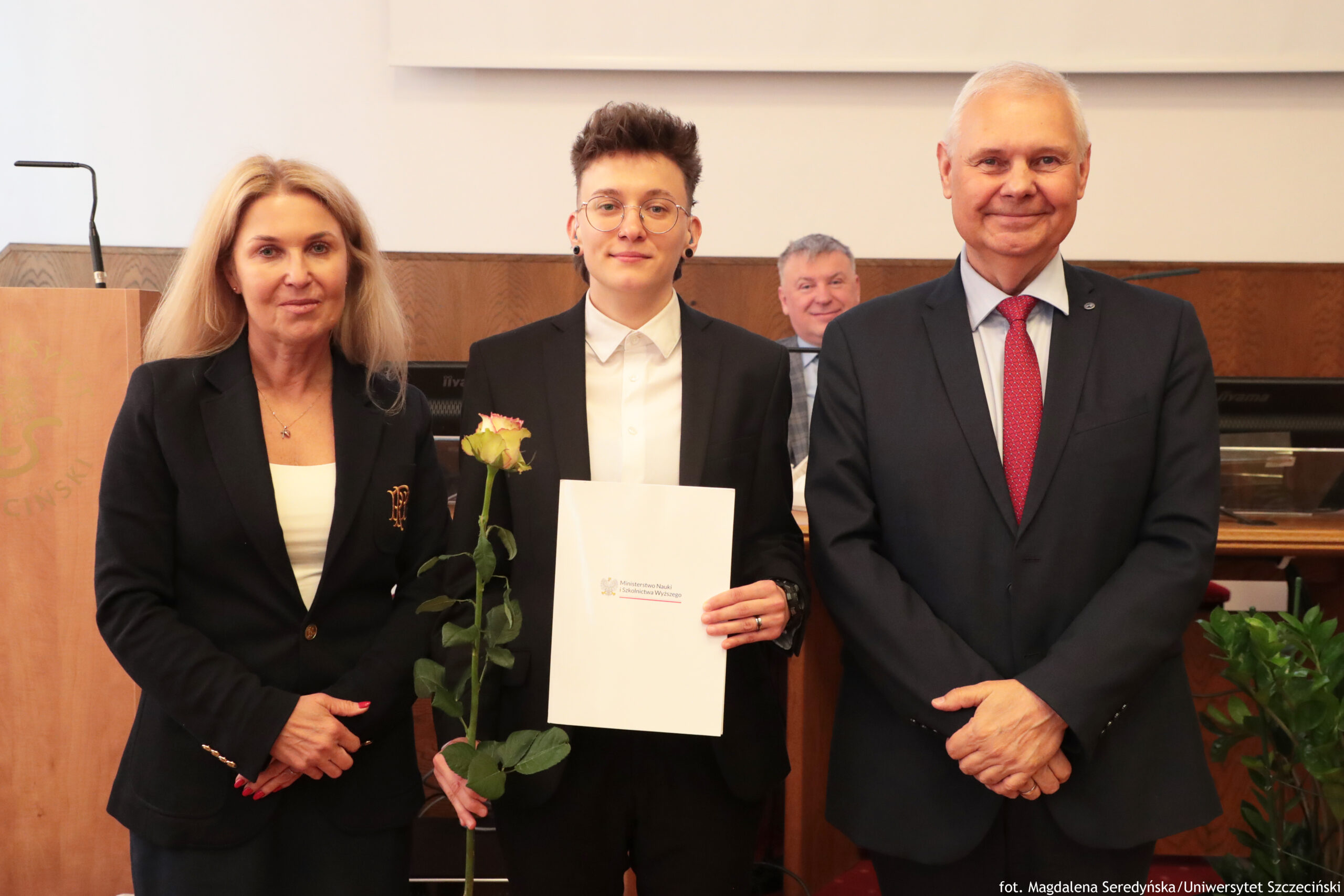 Trzy osoby stoją obok siebie w pomieszczeniu urzędowym lub akademickim. Po lewej kobieta o jasnych, długich, rozpuszczonych włosach, o jasnej karnacji, ubrana w ciemny żakiet i jasny top, z dłońmi złożonymi przed sobą. Pośrodku osoba o krótkich, nastroszonych brązowych włosach i jasnej karnacji, w okularach w cienkich oprawkach, ubrana w białą koszulę i ciemną marynarkę; trzyma pojedynczą, żółtawą różę z zielonymi liśćmi oraz biały sztywny folder lub teczkę z nadrukowanym logo i napisem, częściowo po polsku, wskazującym na instytucję ministerialną. Po prawej mężczyzna o jasnej karnacji i krótkich siwych włosach, w ciemnym garniturze, jasnej koszuli i czerwonym krawacie, z dłońmi splecionymi przed sobą, uśmiechnięty. W tle, na siedząco, widoczny jest starszy mężczyzna w garniturze, uśmiechnięty, za stołem z monitorami komputerowymi. Otoczenie wskazuje na salę konferencyjną lub aulę z drewnianym wykończeniem i mównicą z lewej strony.