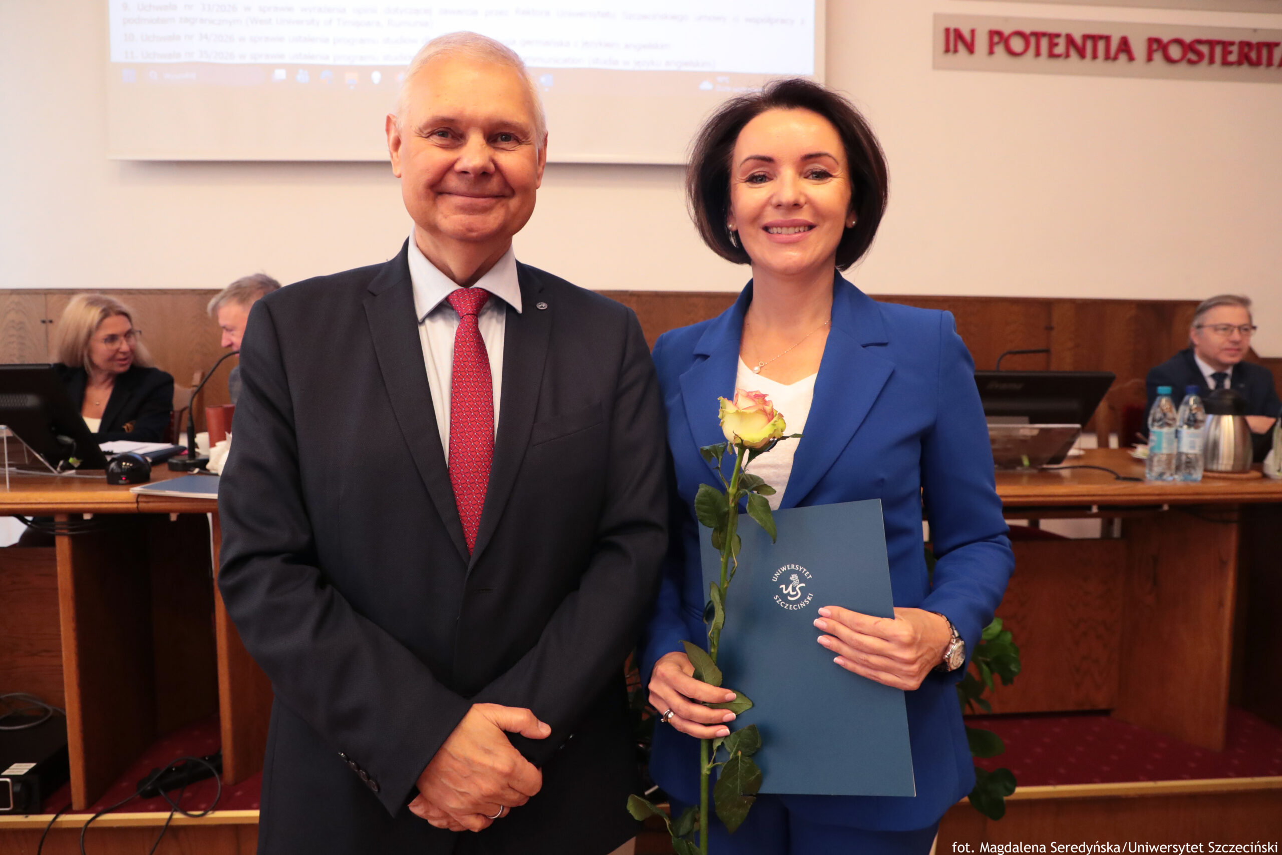 Dwoje dorosłych stojących obok siebie w sali konferencyjnej. Po lewej stronie stoi starszy mężczyzna o jasnej skórze i siwych, krótko ostrzyżonych włosach, ubrany w ciemny garnitur, jasnoniebieską koszulę i czerwony krawat w drobny wzór; trzyma dłonie splecione przed sobą i uśmiecha się. Po prawej znajduje się kobieta o jasnej skórze i ciemnych, krótkich włosach, ubrana w niebieski garnitur i białą bluzkę; uśmiecha się, w lewej ręce trzyma kremowo-żółtą różę, a w prawej niebieską teczkę z białym okrągłym logo i napisem „Uniwersytet Szczeciński”. W tle siedzą przy stołach inni dorośli, widać monitory, mikrofony, butelki z wodą i dzbanki, a nad nimi znajduje się duży ekran projekcyjny.