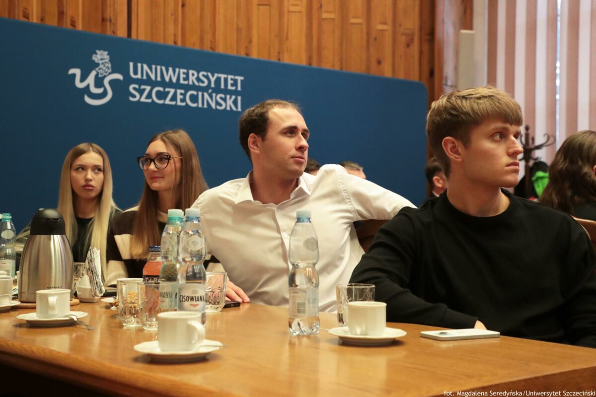 Grupa młodych osób siedzących przy długim stole podczas spotkania w sali z drewnianą ścianą. W tle stoi duża granatowa tablica z białym logo i napisem: „UNIWERSYTET SZCZECIŃSKI”. Na pierwszym planie widać stół z filiżankami na spodkach, szklankami, metalowym dzbankiem termicznym oraz kilkoma butelkami wody. Jedna z butelek ma widoczną etykietę „CISOWIANKA”. Przy stole siedzą m.in. mężczyzna w białej koszuli (na środku) i mężczyzna w czarnym swetrze (po prawej), obaj zwróceni głowami w prawą stronę, jakby słuchali kogoś poza kadrem. Po lewej stronie siedzą dwie młode kobiety; jedna ma długie jasne włosy, druga długie brązowe włosy i duże okulary. W tle po prawej widać wieszak z ubraniami. W prawym dolnym rogu zdjęcia jest mały podpis: „fot. Magdalena Seredyńska/Uniwersytet Szczeciński”.