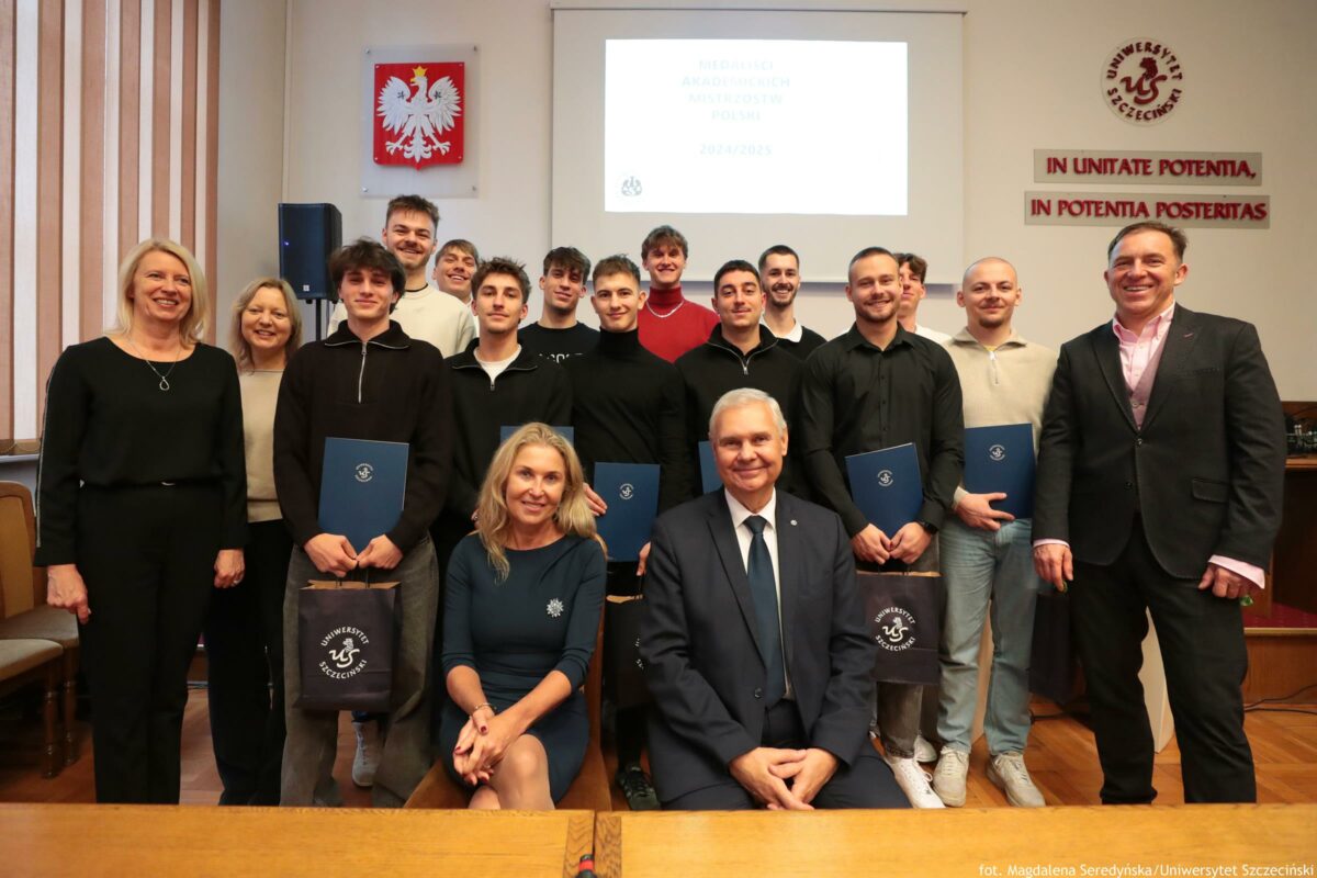 Grupowe zdjęcie w sali uniwersyteckiej z kilkunastoma osobami pozującymi do fotografii, część stojąca w dwóch rzędach i dwie osoby siedzące z przodu. Większość osób to młodzi mężczyźni, kilka osób to kobiety; wszyscy są ubrani w stroje półformalne lub casual (swetry, koszule, marynarki). Kilka osób trzyma granatowe teczki oraz ciemne torby z jasnym logo i napisem „Uniwersytet Szczeciński”. Na ścianie pośrodku, nad grupą, wisi godło Polski (biały orzeł na czerwonym tle). Po prawej stronie ściany znajduje się okrągłe logo uczelni oraz dwa czerwone napisy łacińskie: „IN UNITATE POTENTIA” i „IN POTENTIA POSTERITAS”. Nad grupą jest ekran projekcyjny z bardzo jasnym slajdem; widoczny jest rok „2024/2025”, a reszta tekstu jest zbyt mało czytelna na tym zdjęciu. W prawym dolnym rogu widnieje podpis: „fot. Magdalena Seredyńska/Uniwersytet Szczeciński”.