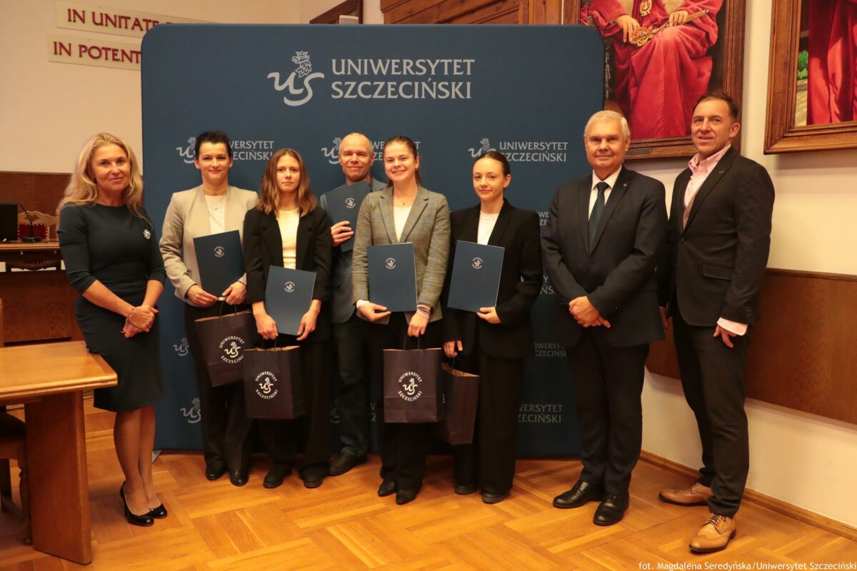 Grupa ośmiu osób stojących w pomieszczeniu przed granatową ścianką z napisem „UNIWERSYTET SZCZECIŃSKI” i logo uczelni. W centrum stoją cztery młode osoby w formalnych strojach, trzymające granatowe teczki z logo oraz torby prezentowe z napisem „Uniwersytet Szczeciński”. Obok nich stoi mężczyzna w średnim wieku w marynarce, również z teczką. Po bokach grupy stoją trzy osoby: po lewej kobieta w ciemnej sukience i kobieta w jasnej marynarce, a po prawej dwóch mężczyzn w garniturach. Tło to sala z drewnianą podłogą; po prawej na ścianie wisi duży obraz w czerwonych barwach. W lewym górnym rogu widać fragmenty łacińskich napisów na ścianie. Na dole po prawej jest podpis: „fot. Magdalena Seredyńska/Uniwersytet Szczeciński”.