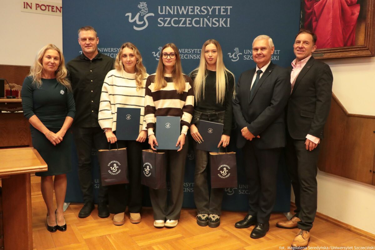 Grupa siedmiu osób stojących w szeregu na tle ścianki z napisem „UNIWERSYTET SZCZECIŃSKI” i logo „US”. Na zdjęciu są cztery kobiety i trzech mężczyzn, wszyscy pozują do fotografii w pomieszczeniu (drewniana podłoga). Trzy kobiety w środku trzymają granatowe teczki/dyplomy z logo oraz czarne torby z napisem i logo „Uniwersytet Szczeciński”. Od lewej: kobieta w dopasowanej ciemnozielonej sukience, blond włosy. Obok mężczyzna w czarnej koszuli. Następnie kobieta w biało-czarnym swetrze w poziome pasy. Kolejna kobieta w podobnym swetrze w pasy, ma okulary i długie proste włosy. Dalej kobieta z bardzo długimi blond włosami, w ciemnym stroju. Następnie starszy mężczyzna w garniturze i krawacie. Po prawej mężczyzna w ciemnym garniturze z jasnoróżową koszulą. W lewym dolnym rogu widać fragment drewnianego stołu. W prawym górnym rogu widać część obrazu w ramie z czerwonym elementem. Widoczny tekst: „UNIWERSYTET SZCZECIŃSKI”, logo „US”, na torbach „UNIWERSYTET SZCZECIŃSKI”. W prawym dolnym rogu drobny podpis: „fot. Magdalena Seredyńska/Uniwersytet Szczeciński”.