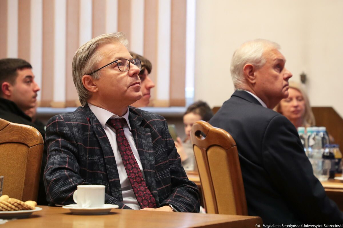Mężczyzna w garniturze w kratę, w okularach, siedzący przy stole w sali konferencyjnej. Ma jasną skórę, siwe włosy zaczesane do tyłu i czerwony krawat we wzór. Jest zwrócony bokiem i ma uniesioną głowę, jakby słuchał kogoś poza kadrem. Na stole przed nim stoi biała filiżanka na spodku, a z lewej strony częściowo widać talerzyk z przekąskami. Po prawej stronie kadru siedzi drugi mężczyzna o jasnej skórze, z siwymi włosami, w ciemnym garniturze, również zwrócony bokiem. W tle znajduje się kilka innych osób siedzących przy stołach oraz pionowe żaluzje w oknie. W prawym dolnym rogu widoczny jest napis: „fot. Magdalena Seredyńska/Uniwersytet Szczeciński”.
