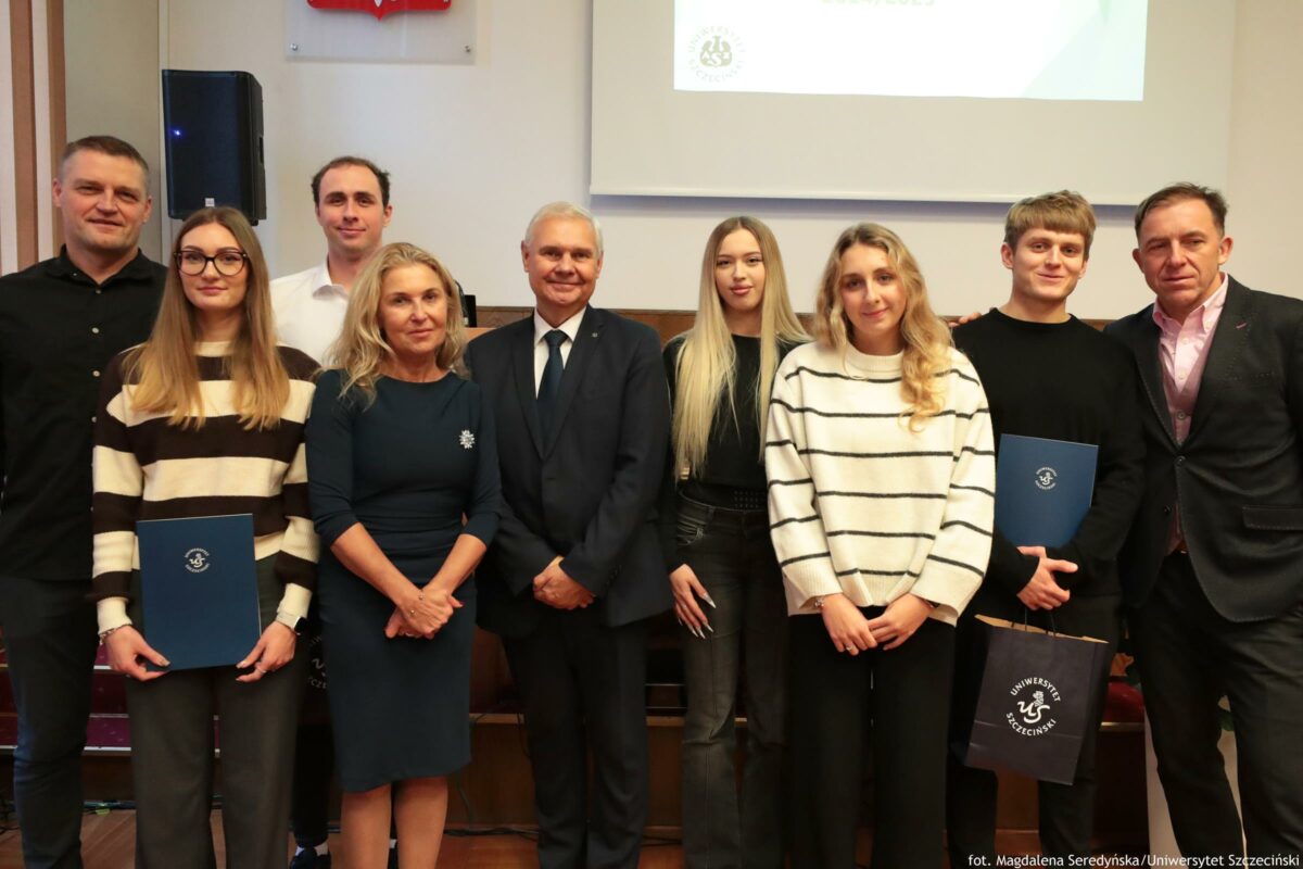 Grupowe zdjęcie dziewięciu osób stojących w sali wykładowej, pozujących do fotografii. Widać pięć osób w pierwszym rzędzie i cztery osoby w drugim rzędzie. Pośrodku stoi starszy mężczyzna o siwych włosach, w ciemnym garniturze i krawacie. Po jego lewej stronie stoi kobieta w granatowej sukience, blond włosy do ramion. Po jego prawej stronie stoi młoda kobieta z długimi prostymi blond włosami, w czarnym stroju. Z przodu po lewej jest młoda kobieta z długimi jasnobrązowymi włosami i okularami, w swetrze w szerokie poziome pasy jasne i ciemne; trzyma granatową teczkę z okrągłym znakiem. Z przodu po prawej jest młoda kobieta z falowanymi blond włosami, w białym swetrze w czarne poziome paski. Po prawej stronie, w drugim rzędzie, stoi młody mężczyzna w czarnym swetrze; trzyma granatową teczkę z okrągłym znakiem oraz torbę z napisem „Uniwersytet Szczeciński”. Skrajnie po prawej stoi mężczyzna w ciemnej marynarce i jasnoróżowej koszuli. Skrajnie po lewej stoi mężczyzna w czarnej koszuli. Za kobietą w okularach stoi mężczyzna w białej koszuli. W tle widać głośnik, fragment ekranu/projektora z niewyraźnym slajdem oraz godło Polski nad ekranem. W prawym dolnym rogu jest podpis fotograficzny: „fot. Magdalena Seredyńska/Uniwersytet Szczeciński”.