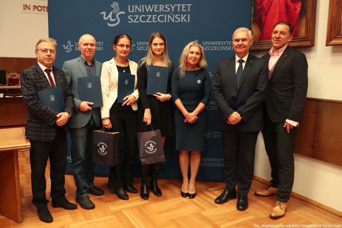 Grupa siedmiu osób stojących w rzędzie na sali, na tle granatowej ścianki z dużym napisem „UNIWERSYTET SZCZECIŃSKI” i logotypem uczelni. To zdjęcie z uroczystości o formalnym charakterze. Od lewej do prawej: Mężczyzna w okularach, w ciemnej marynarce w kratę, białej koszuli i krawacie; trzyma granatową teczkę z logo. Mężczyzna w jasnoniebieskiej marynarce i dżinsach; trzyma podobną teczkę. Kobieta w jasnej (kremowej) marynarce i ciemnych spodniach, w okularach; trzyma teczkę i ma torbę papierową z logo uczelni. Młoda kobieta z długimi jasnymi włosami, w czarnym stroju; trzyma teczkę i ma torbę papierową z logo uczelni. Kobieta z jasnymi włosami w sukience w kolorze ciemnym (granatowym), z ozdobą przypiętą na piersi; ręce splecione z przodu. Mężczyzna w ciemnym garniturze, białej koszuli i krawacie; stoi z rękami złożonymi z przodu. Mężczyzna w ciemnym garniturze i jasnej koszuli; stoi po prawej stronie. Na dole zdjęcia widać drewnianą podłogę. Na prawym brzegu jest fragment obrazu w ramie z czerwonym elementem. W prawym dolnym rogu widoczny podpis: „fot. Magdalena Seredyńska / Uniwersytet Szczeciński”.