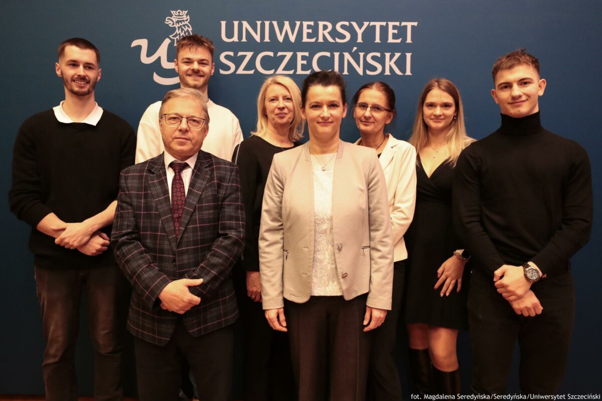Grupowe zdjęcie ośmiu osób stojących w dwóch rzędach na tle granatowej ścianki z dużym białym napisem „UNIWERSYTET SZCZECIŃSKI” oraz białym logo po lewej stronie. W pierwszym rzędzie stoją dwie osoby: po lewej mężczyzna w okularach, w ciemnej kraciastej marynarce, białej koszuli i krawacie; na środku kobieta z ciemnymi włosami upiętymi do góry, w jasnej marynarce i jasnej bluzce. W drugim rzędzie stoi sześć osób. Od lewej: mężczyzna z krótkimi ciemnymi włosami i zarostem, w czarnym swetrze na białym kołnierzyku; obok wysoki mężczyzna z jasnobrązowymi włosami w białej koszuli; dalej blondynka w czarnej bluzce; kobieta w okularach, z ciemnymi włosami związanymi, w jasnej marynarce; młoda kobieta z długimi jasnymi włosami w czarnej sukience; po prawej mężczyzna z krótkimi włosami w czarnym golfie, z zegarkiem na ręku. W prawym dolnym rogu jest mały podpis: „fot. Magdalena Seredyńska/Seredyńska/Uniwersytet Szczeciński”.