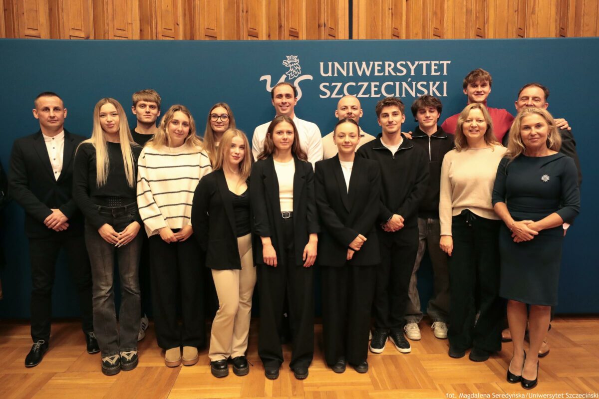 Grupowe zdjęcie kilkunastu osób pozujących na tle ścianki z napisem „UNIWERSYTET SZCZECIŃSKI” oraz białym logo z gryfem. Osoby stoją w dwóch rzędach, w większości w strojach półformalnych lub formalnych. W pierwszym rzędzie widać kilka młodych kobiet ubranych głównie na czarno (marynarki, spodnie), jedna ma jasne spodnie i czarną górę. Po prawej stronie stoją dwie dorosłe kobiety; jedna w jasnym swetrze i ciemnych spodniach, druga w granatowej sukience z broszką. W drugim rzędzie stoją młodzi mężczyźni oraz dwaj dorośli mężczyźni; jeden z nich jest w białej koszuli, inny jest łysy z zarostem. Po lewej stronie stoi mężczyzna w ciemnym garniturze i białej koszuli oraz młoda kobieta z długimi, jasnymi włosami w czarnym stroju. Po prawej w tle widać wysokiego młodego mężczyznę w czerwonej bluzie. Na dole, przy prawej krawędzi, znajduje się mały podpis: „fot. Magdalena Seredyńska / Uniwersytet Szczeciński”.