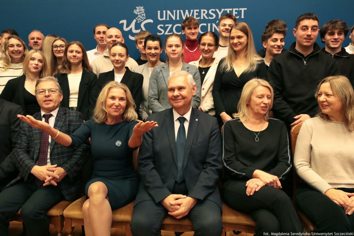 Grupowe zdjęcie kilkunastu osób pozujących na tle niebieskiej ścianki z napisem „UNIWERSYTET SZCZECIŃSKI” oraz białym logo uczelni. W pierwszym rzędzie na krzesłach siedzi pięć osób: mężczyzna w okularach w kraciastej marynarce, kobieta w ciemnej sukience z broszką i uniesionymi dłońmi w geście prezentowania, starszy mężczyzna w granatowym garniturze i krawacie, kobieta w czarnym stroju z jasnymi włosami oraz kobieta w jasnym swetrze. Za nimi stoi duża grupa młodych osób (kobiet i mężczyzn) w strojach półformalnych: marynarki, koszule, swetry; większość uśmiechnięta i skierowana w stronę aparatu. W prawym dolnym rogu widoczny jest podpis: „fot. Magdalena Seredyńska / Uniwersytet Szczeciński”.