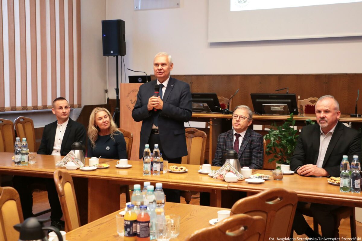 Sala konferencyjna z długimi drewnianymi stołami, przy których siedzi kilka osób, a jedna osoba stoi i przemawia z mikrofonem. Na środku stoi starszy mężczyzna z siwymi włosami w ciemnym garniturze i białej koszuli. Trzyma mikrofon obiema rękami i jest zwrócony przodem do zebranych. Przy stole, od lewej strony, siedzi mężczyzna w czarnej marynarce i białej koszuli. Obok niego siedzi blond kobieta w ciemnej bluzce. Po prawej stronie stojącego mężczyzny siedzi mężczyzna w okularach, w marynarce w kratę i krawacie. Najbardziej po prawej siedzi mężczyzna z krótkimi siwymi włosami i zarostem, w ciemnej marynarce i jasnej koszuli. Na stołach stoją butelki wody, szklanki, filiżanki, talerzyki i półmiski z drobnymi przekąskami; widać też dzbanki/termosy oraz butelki napojów. W tle jest mównica z częściowo widocznym napisem „Uniwer…”, sprzęt nagłośnieniowy, monitory na stanowisku oraz fragment ekranu/projektora u góry. Po lewej stronie są pionowe żaluzje w oknie. W prawym dolnym rogu widoczny jest podpis: „fot. Magdalena Seredyńska/Uniwersytet Szczeciński”.