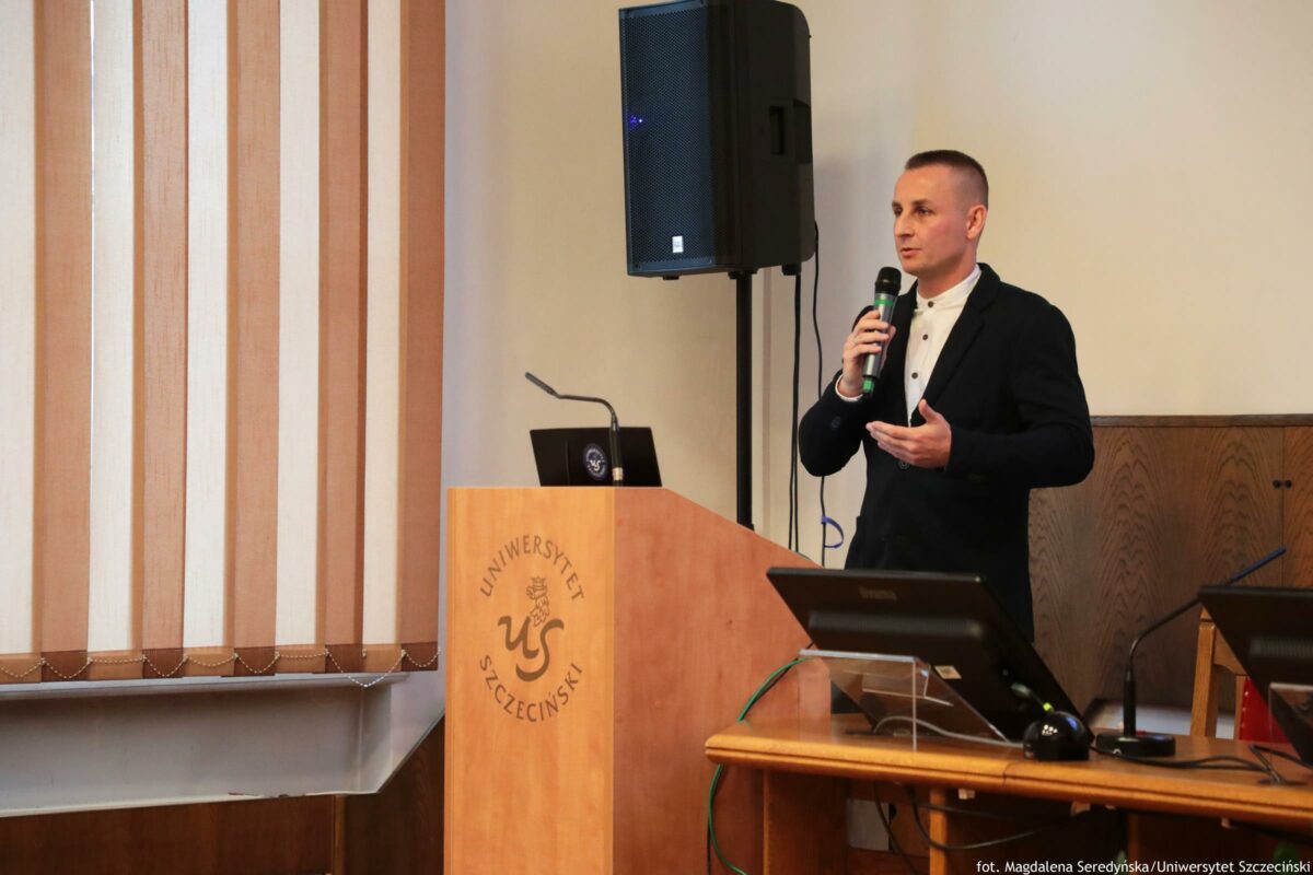 Mężczyzna w ciemnym garniturze przemawiający z mikrofonem przy mównicy z napisem „UNIWERSYTET SZCZECIŃSKI”. Ma krótkie, ciemne włosy i stoi po prawej stronie kadru, zwrócony lekko w lewo, z jedną dłonią uniesioną w geście podczas mówienia. Na mównicy stoi otwarty laptop i cienki mikrofon na giętkim wysięgniku. Za mównicą znajduje się duży czarny głośnik na statywie. Po lewej stronie są pionowe żaluzje w odcieniach beżu i brązu. Na pierwszym planie widać stół z monitorem, przewodami i sprzętem konferencyjnym. W prawym dolnym rogu widnieje podpis: „fot. Magdalena Seredyńska/Uniwersytet Szczeciński”.