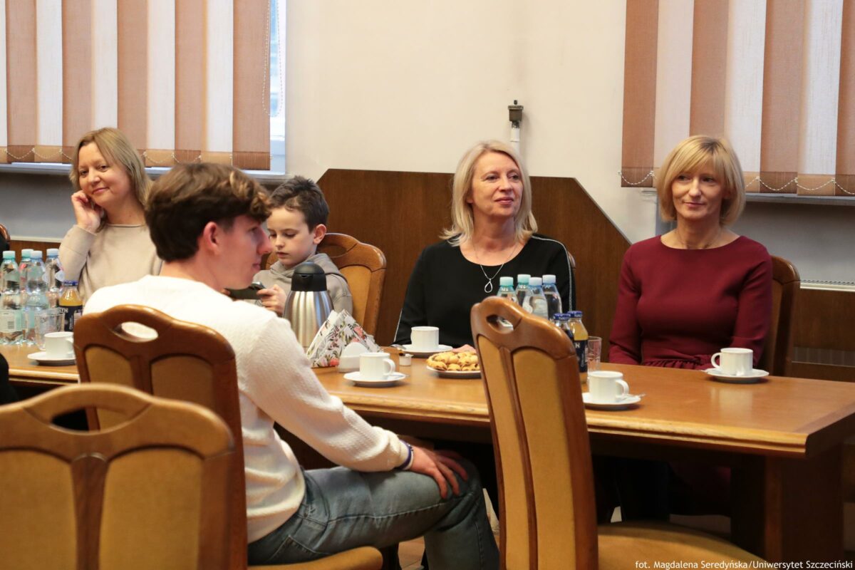 Grupa osób siedząca przy drewnianym stole w sali z pionowymi żaluzjami w oknach. Przy stole siedzą cztery osoby dorosłe i jedno dziecko. Na pierwszym planie po lewej widać chłopca lub młodego mężczyznę z ciemnymi włosami w białym swetrze i niebieskich dżinsach, odwróconego bokiem. Za nim, po lewej stronie stołu, siedzi kobieta w jasnym swetrze, opierająca głowę na dłoni. Obok niej siedzi dziecko z krótkimi, ciemnymi włosami. Pośrodku, dalej przy stole, siedzi kobieta o jasnych włosach w czarnej bluzce. Po prawej stronie siedzi kobieta o jasnych włosach w bordowej sukience. Na stole stoją białe filiżanki ze spodkami, dzbanek termosowy, butelki wody i soku oraz talerz z ciastkami. W prawym dolnym rogu jest podpis: „fot. Magdalena Seredyńska/Uniwersytet Szczeciński”.