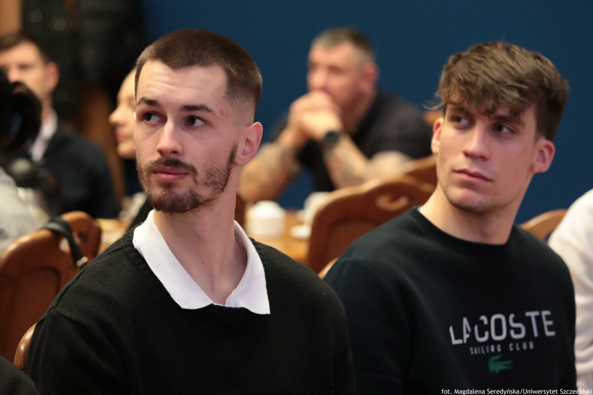 Dwóch młodych mężczyzn siedzących na krzesłach na sali spotkania lub wykładu. Na pierwszym planie po lewej stronie jest mężczyzna o jasnej karnacji, z bardzo krótko ostrzyżonymi włosami i krótką brodą; ma na sobie czarną bluzę z białym kołnierzykiem i jest zwrócony głową w lewo. Po prawej stronie siedzi drugi mężczyzna o jasnej karnacji, z brązowymi, lekko potarganymi włosami; ma czarną bluzę z napisem „LACOSTE” i również patrzy w lewo. W tle są rozmyte sylwetki innych osób siedzących przy stołach oraz drewniane oparcia krzeseł. W prawym dolnym rogu widoczny jest mały podpis: „fot. Magdalena Seredyńska/Uniwersytet Szczeciński”.”