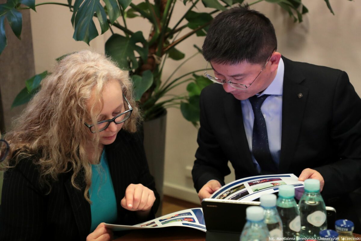 Dwie osoby siedzące przy stole i przeglądające materiały w formie broszur lub wydruków. Po lewej stronie jest kobieta o jasnej skórze, z długimi, kręconymi blond włosami i okularami. Ma na sobie turkusową bluzkę i czarną narzutkę lub żakiet. Patrzy w dół na otwartą broszurę. Po prawej stronie jest mężczyzna o jasnej skórze, z krótkimi ciemnymi włosami i okularami. Ma na sobie czarny garnitur, jasną koszulę i ciemny krawat. Trzyma otwarty katalog lub broszurę i też patrzy w dół.