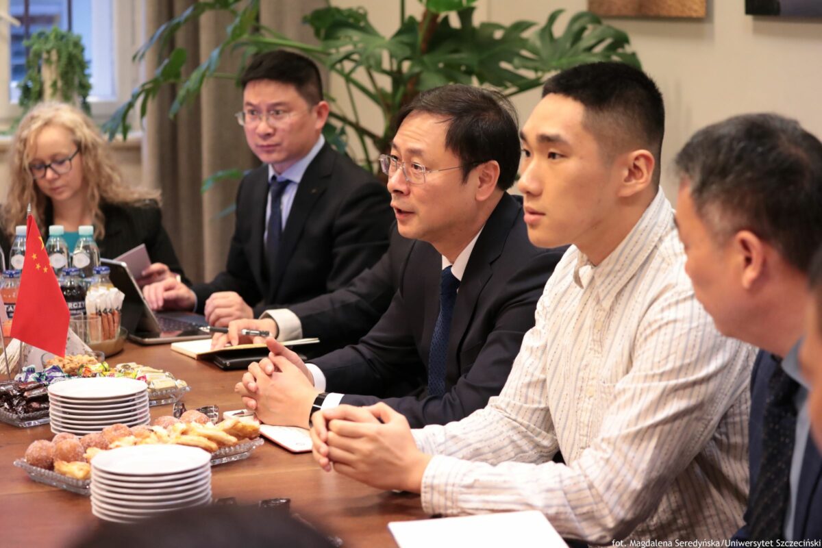 Grupa osób siedzących przy stole konferencyjnym podczas spotkania. Widać cztery osoby na pierwszym planie: trzech mężczyzn i jedną kobietę w tle po lewej; wszyscy w stroju biznesowym, jeden z mężczyzn po prawej ma jasną, pasiastą koszulę bez marynarki. Na stole stoją talerze, ciastka i słodycze na paterach, stosy białych talerzyków, butelki wody oraz mała czerwona flaga Chin na stojaku. W tle są duże zielone rośliny doniczkowe i fragmenty obrazów na ścianie. W prawym dolnym rogu jest napis: „fot. Magdalena Seredyńska/Uniwersytet Szczeciński”.