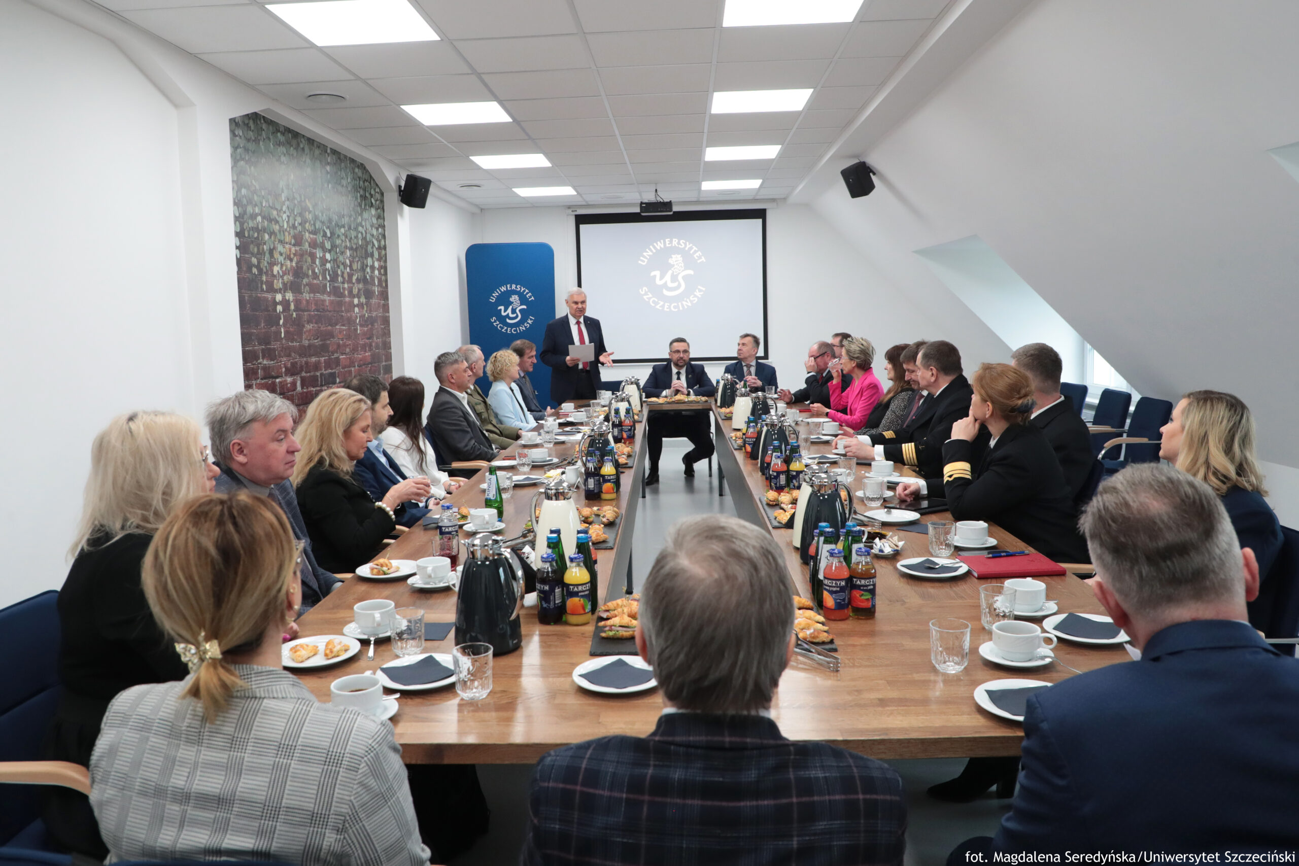 Sala konferencyjna z długim stołem i grupą osób w formalnych strojach podczas spotkania. Osoby siedzą po obu stronach stołu, a na jego końcu stoi mężczyzna w garniturze, wyglądający na prowadzącego lub przemawiającego. Na ścianie z tyłu wisi ekran z logo i napisem „Uniwersytet Szczeciński”, obok stoi niebieski roll-up z tym samym logo. Na stole znajdują się filiżanki ze spodkami, szklanki, dzbanki termiczne, butelki z napojami, talerzyki z przekąskami (w tym drobne kanapki i ciastka) oraz serwetki. Pomieszczenie ma biały sufit z panelami i jasnym oświetleniem; po prawej stronie są skośne ściany z oknami dachowymi. W prawym dolnym rogu widnieje podpis: „fot. Magdalena Seredyńska/Uniwersytet Szczeciński”.